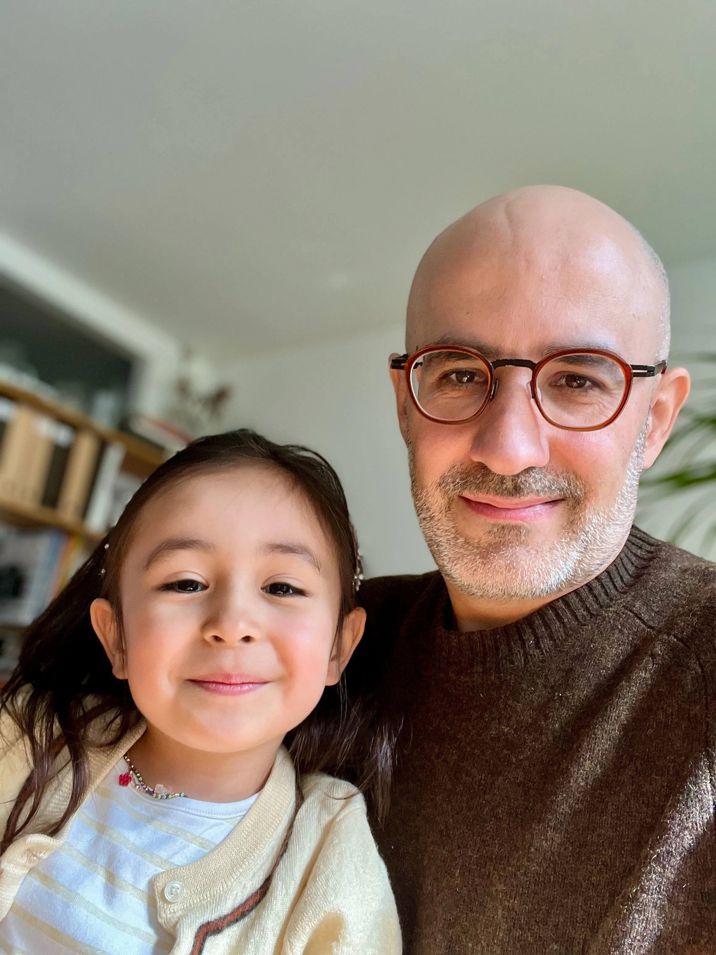 Un homme et une jeune fille prennent un selfie ensemble à l'intérieur d'une maison, la jeune fille sourit et porte un pull ivoire, l'homme porte des lunettes rondes et un pull marron, un fond flou d'étagères et de plantes
