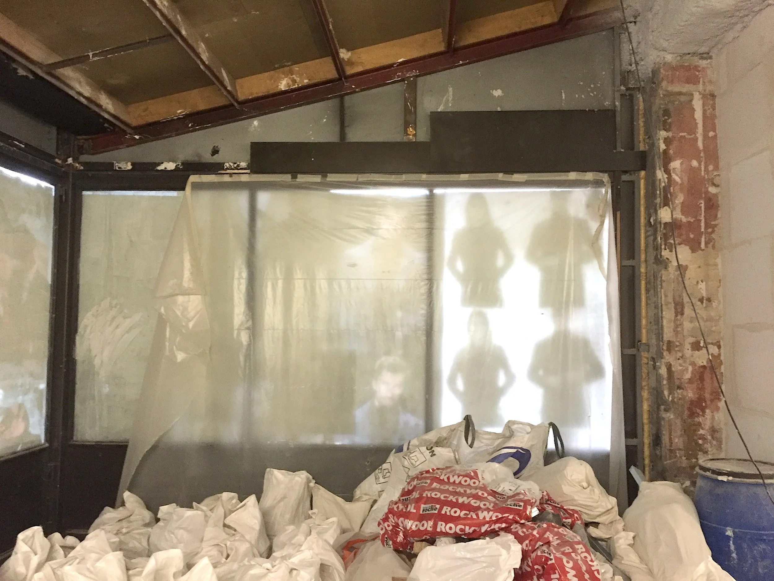 Une pièce en travaux avec des sacs de matériaux empilés devant une cloison en verre partiellement installée. La zone est en rénovation, avec des murs en briques et un plafond en cours d'aménagement.
