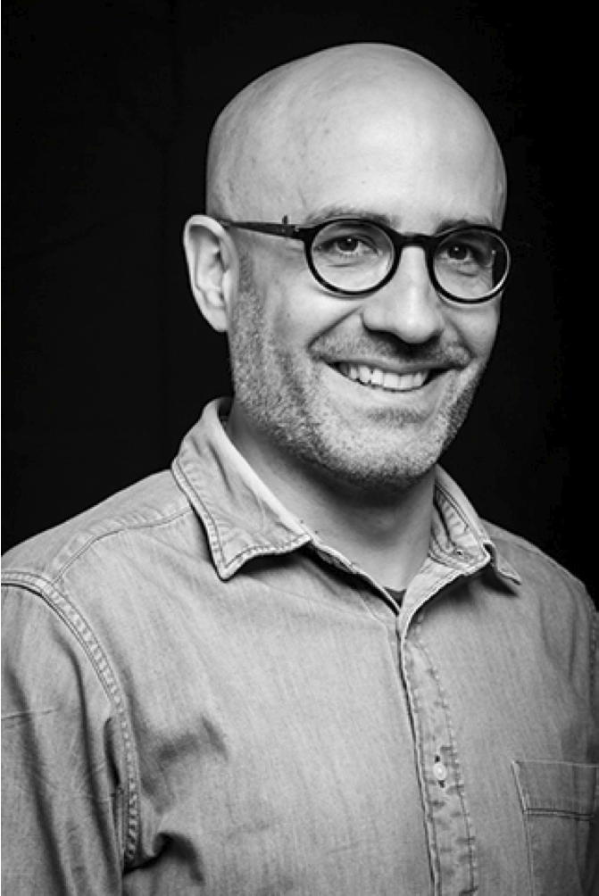 Un portrait en noir et blanc d'un homme souriant, portant des lunettes rondes et une chemise en denim, avec un fond sombre.