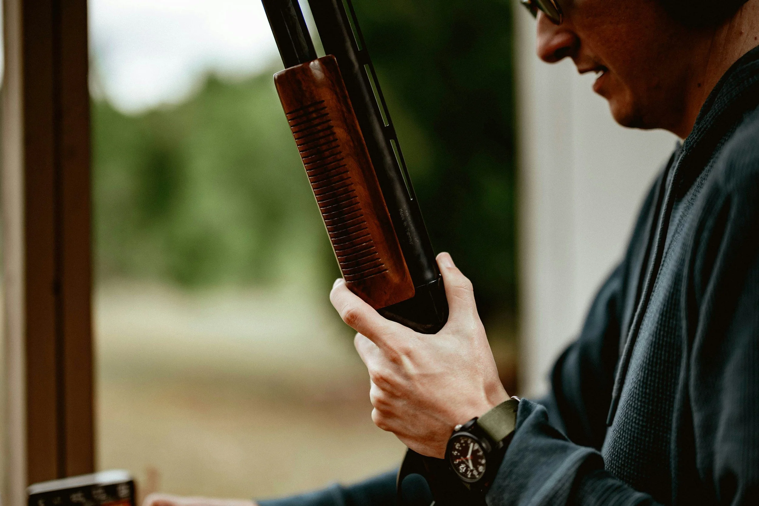 A person with light skin holding a shotgun, standing indoors near a window with a green outdoor view. The person is wearing a dark shirt, a watch, and glasses.