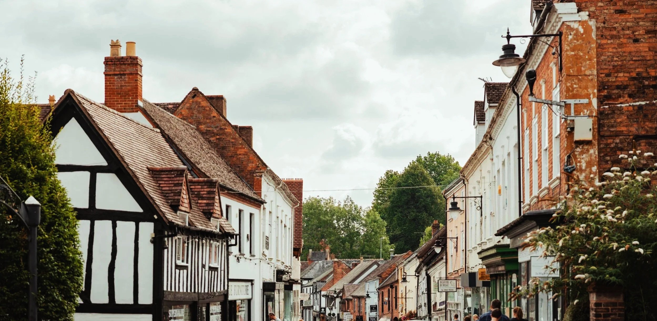 A street scene with historic brick and half-timbered buildings, shops, and pedestrians under a cloudy sky.
