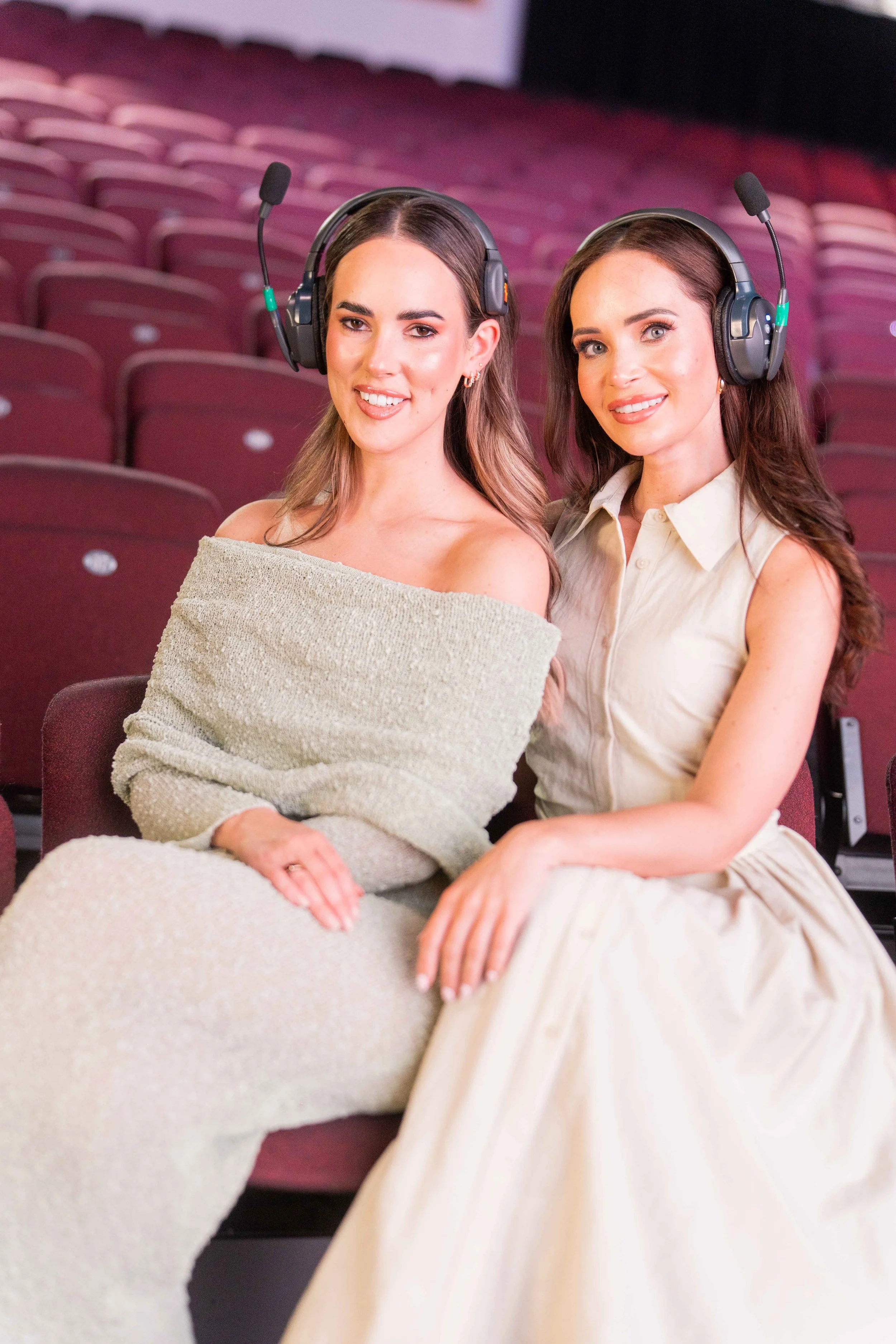 Two women sitting side by side in theater seats, wearing headsets with microphones, smiling at the camera.
