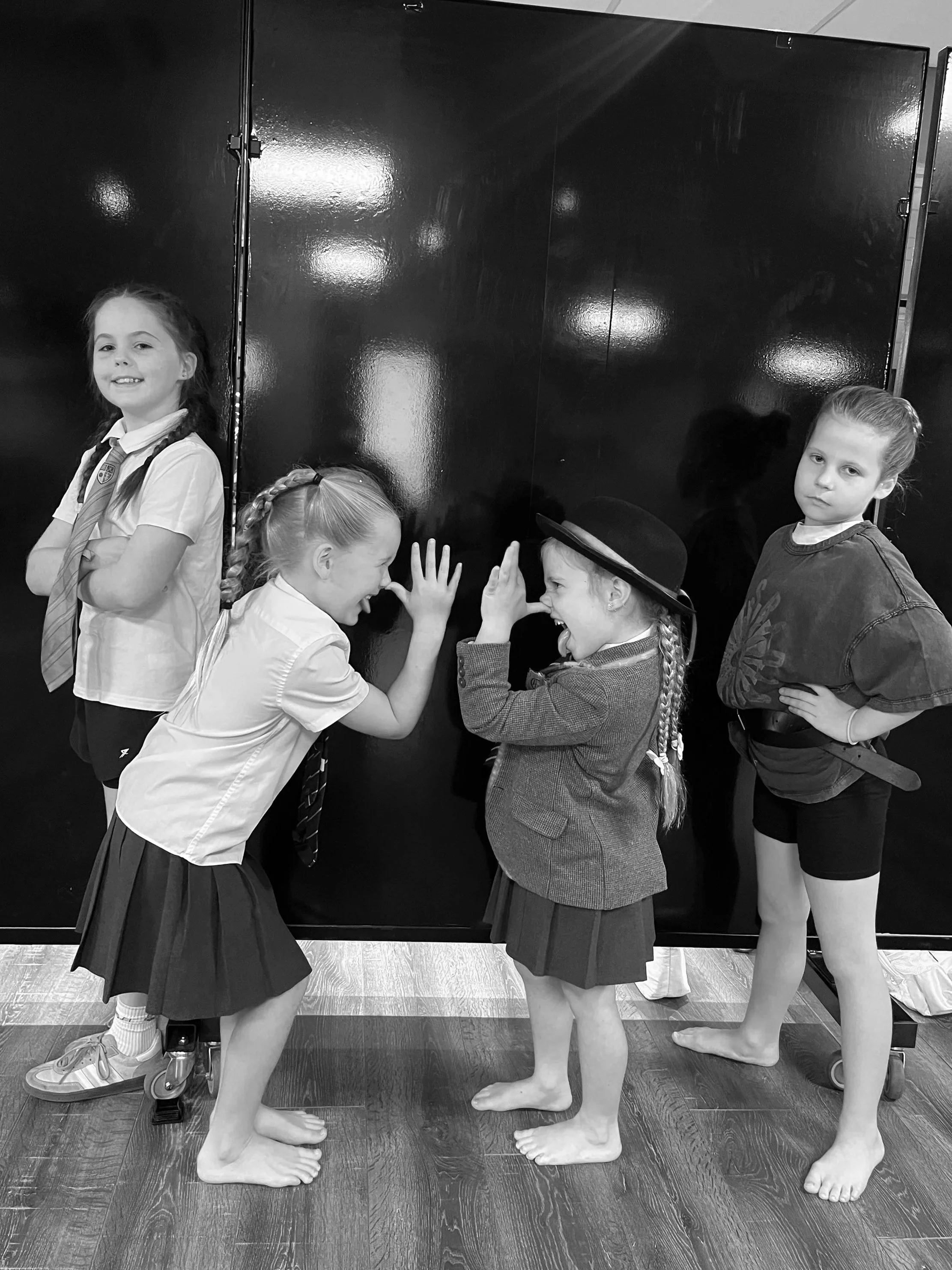 Four young girls in school uniforms and casual clothing standing and facing a pair of mannequins with human-like limbs, engaged in a friendly arm-wrestling match. One girl is on the left, wearing a white shirt and a dark skirt, with a braid, and another on the right, dressed in a blazer and skirt, also with a braid. Two mannequins are present, one on the left, partially visible, and the other on the right, wearing a hat and a sweater. The scene is indoors with a shiny black wall in the background.
