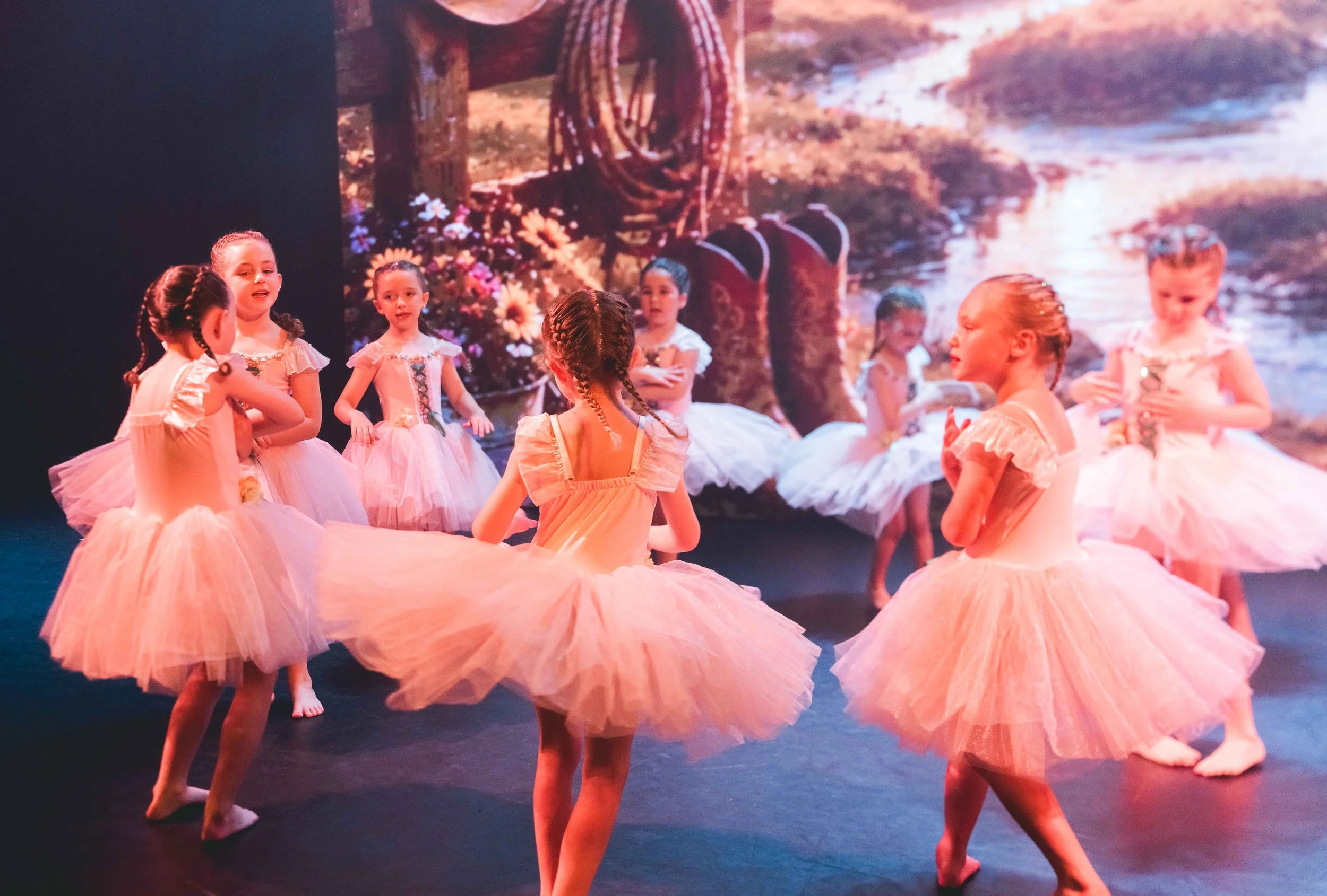 Group of young ballerinas dancing in pink tutus during a performance with a scenic backdrop of a river and mountains.