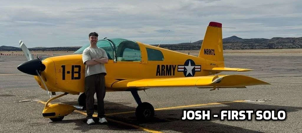 A young man in casual clothing standing with arms crossed next to a yellow military aircraft on an airstrip with mountains in the background.