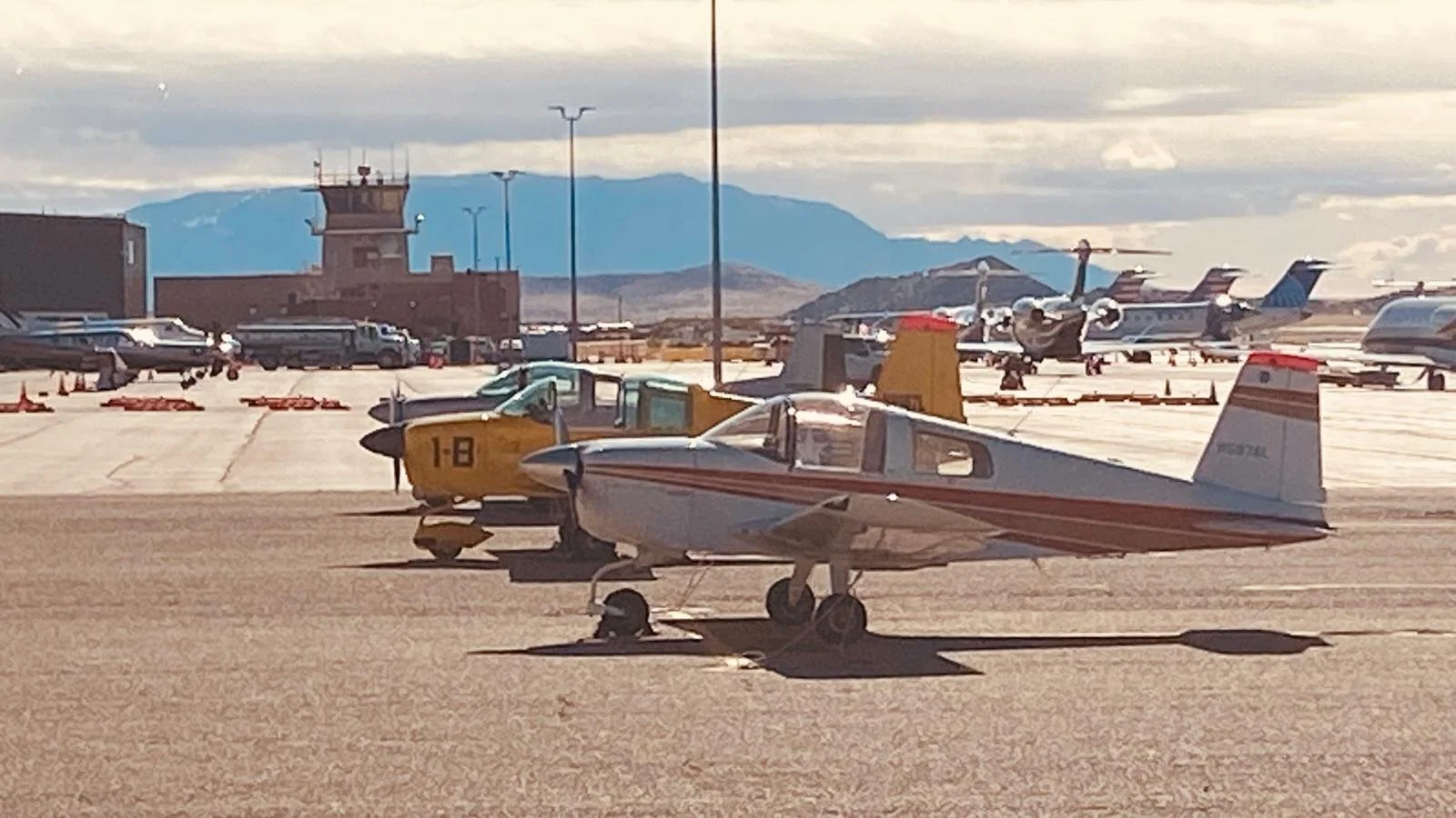 Small private airplanes parked on an airport tarmac with a control tower and mountains in the background during daytime.