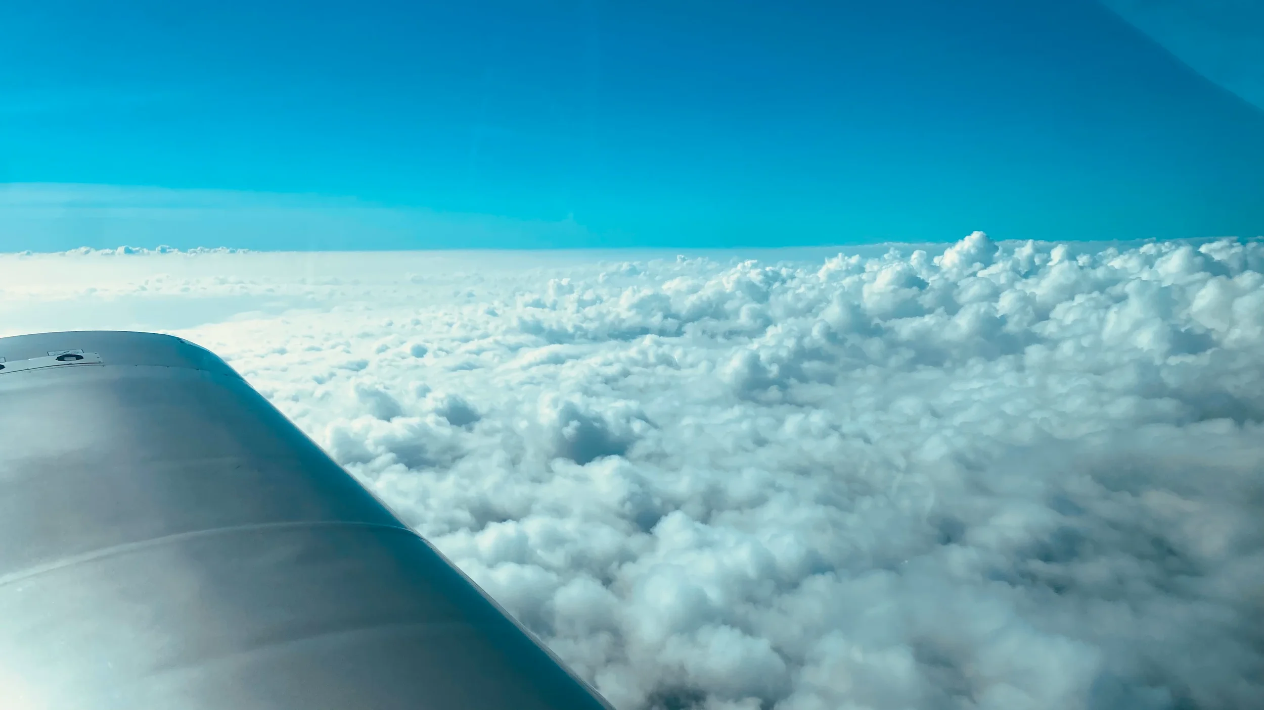 View from an airplane window showing a white airplane wing in the foreground and a vast expanse of fluffy white clouds below, with a blue sky above.
