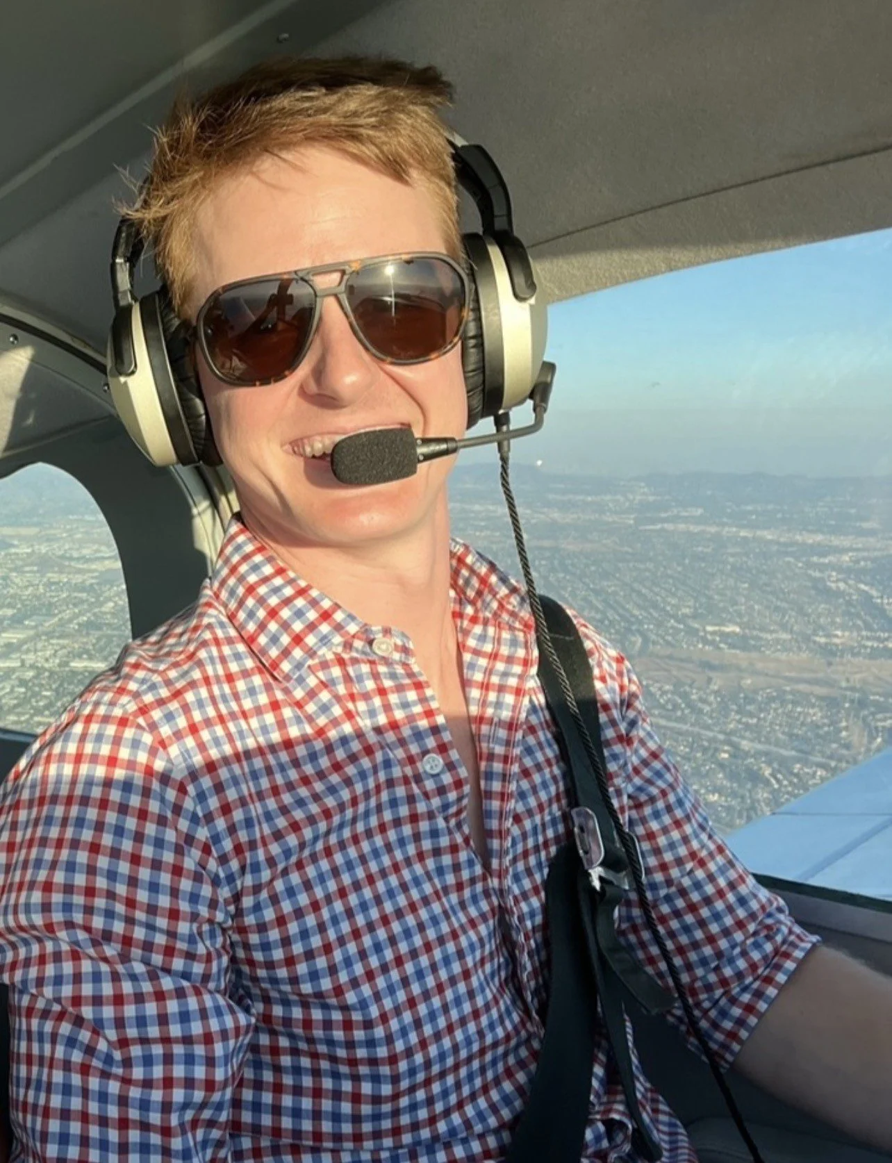 Woman flying a small airplane, wearing sunglasses, a headset with microphone, and a red checkered shirt, with a view of the sky and cityscape outside the window.