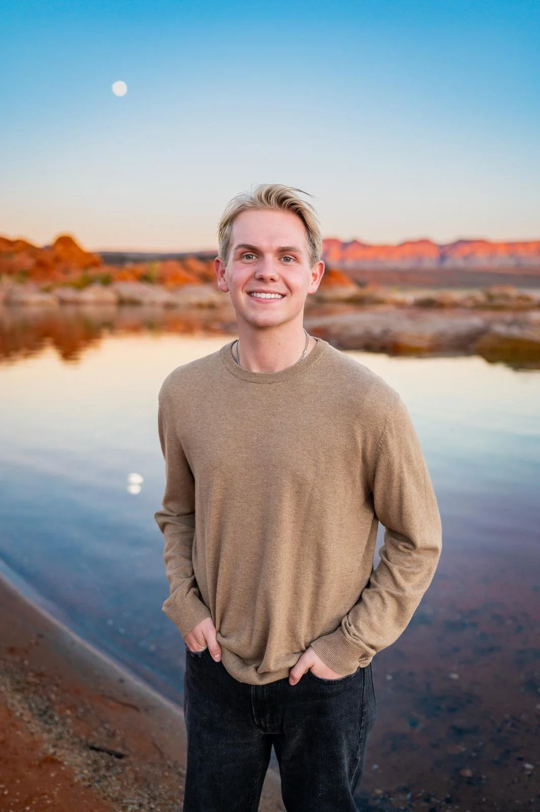 A young man standing outdoors by a body of water during sunset, with mountains and a clear sky in the background.