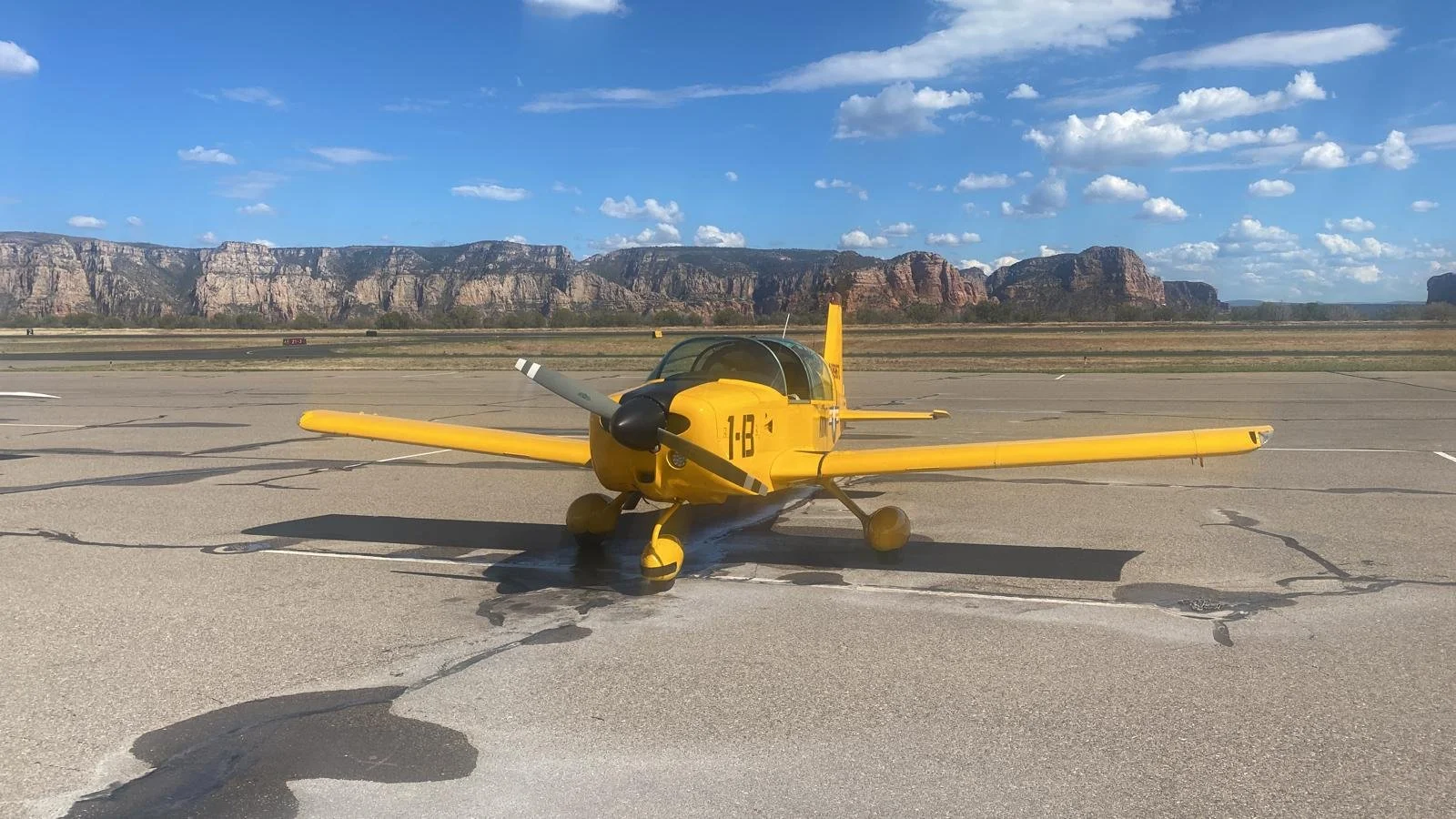 Yellow small aircraft parked on tarmac with mountains in the background and blue sky with scattered clouds.