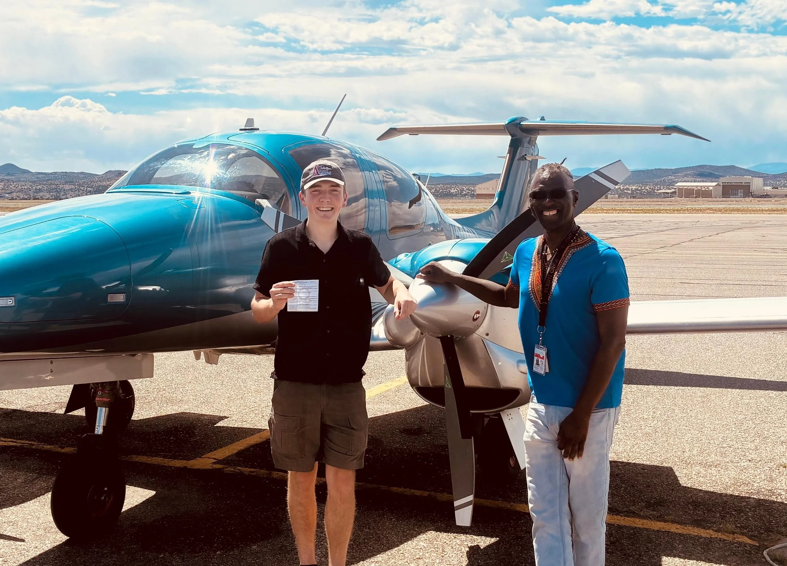 Two men standing next to a blue light aircraft on a runway, with mountains and a building in the background. One man is holding a document, the other is smiling and wearing sunglasses.