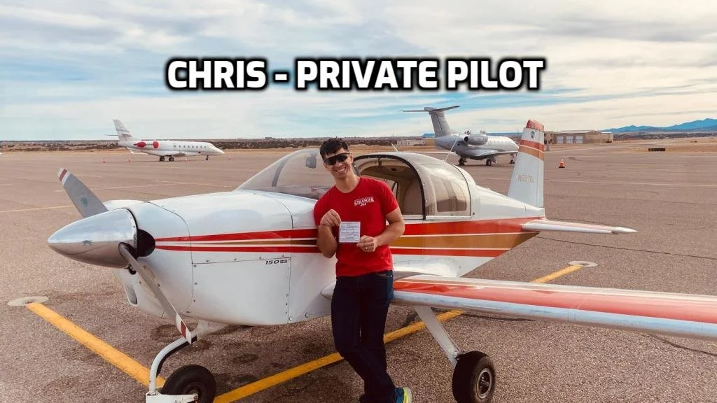 A man in a red shirt standing next to a small propeller airplane on a runway, holding a certificate, with two larger planes in the background, under a cloudy sky.