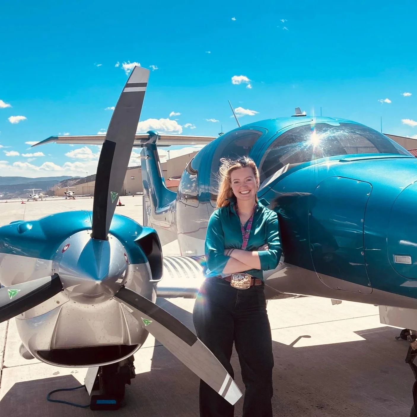 A woman standing in front of a blue airplane on a sunny day, smiling with her arms crossed.