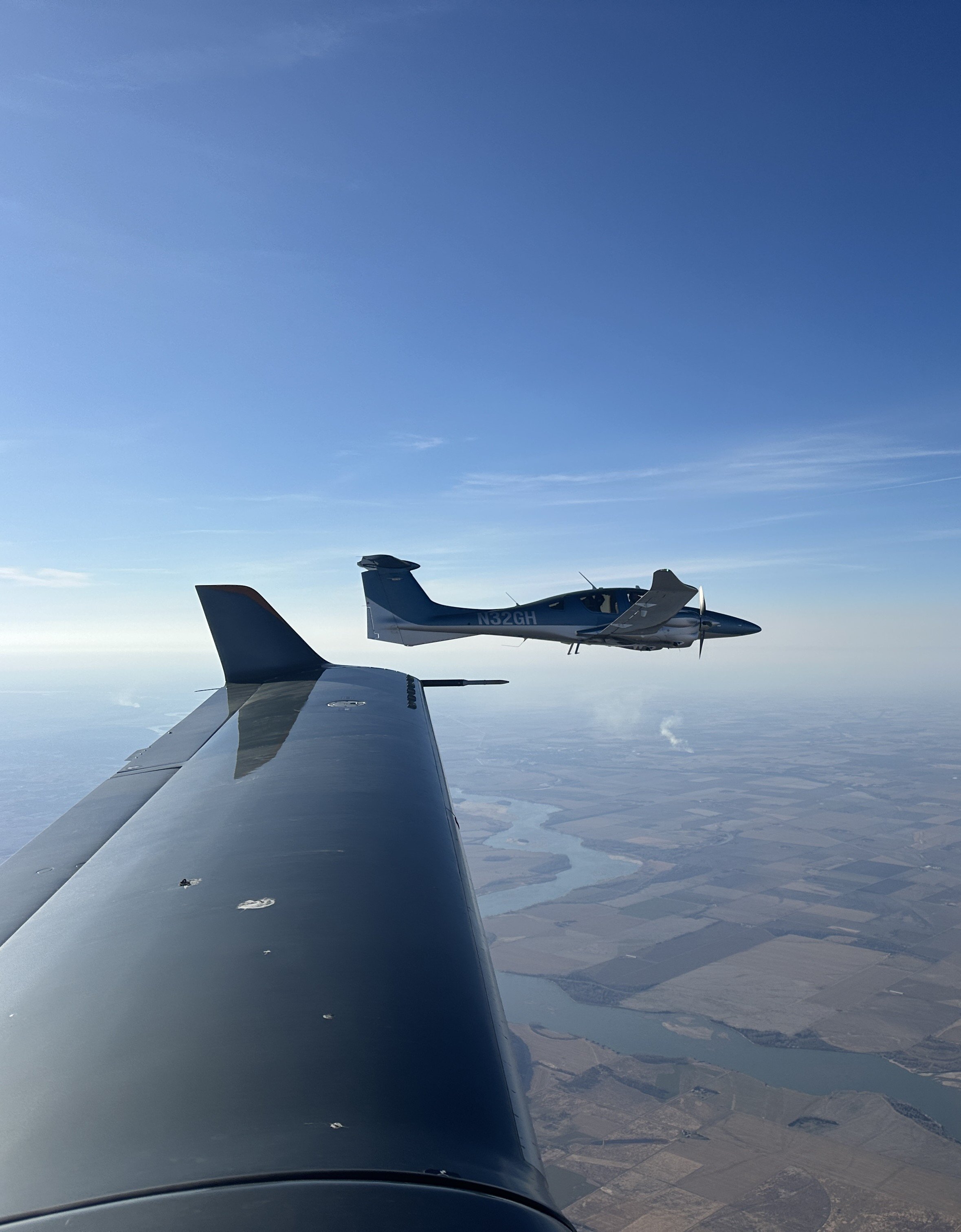 View from airplane wing showing two small aircraft flying in the sky above a landscape of fields and bodies of water, with a clear blue sky.
