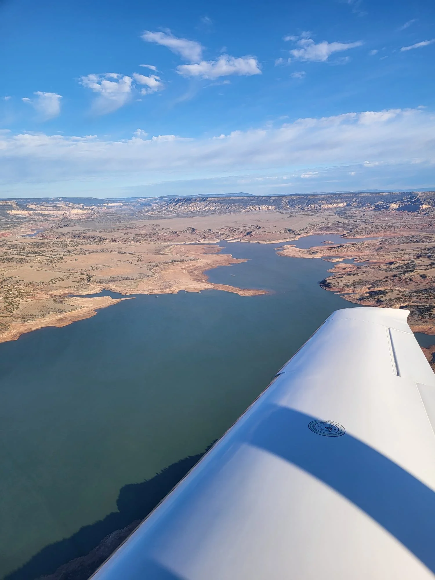 Aerial view of a river and arid landscape taken from an airplane window, with part of the airplane wing visible in the lower right corner, under a partly cloudy blue sky.