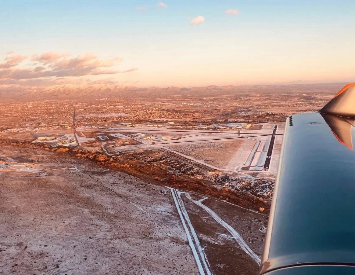 Aerial view from an airplane showing a snowy landscape with roads, airport runways, and a city in the distance during sunset.