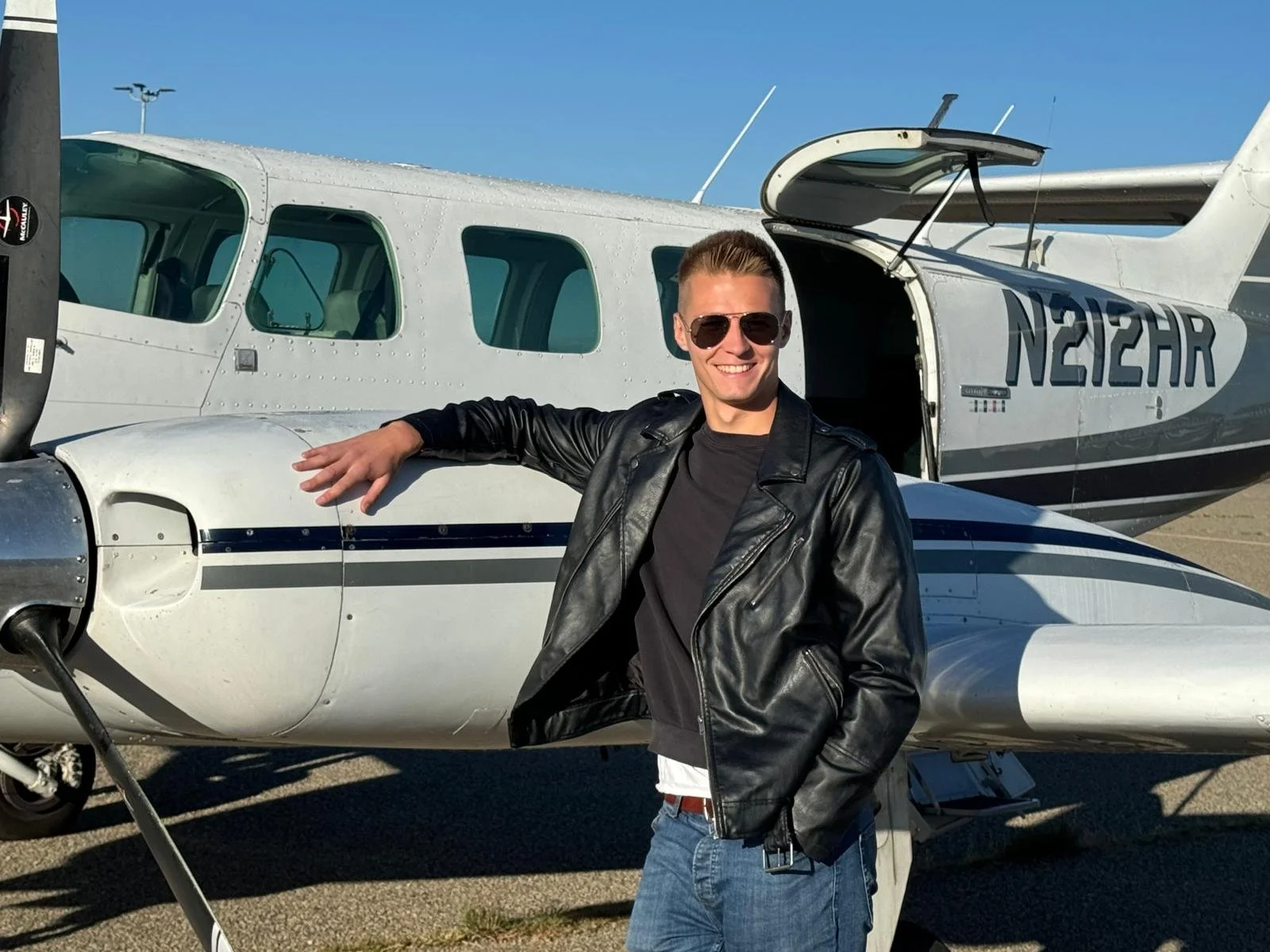 A young man in sunglasses and a black leather jacket smiling, standing in front of a small white airplane on a sunny day.