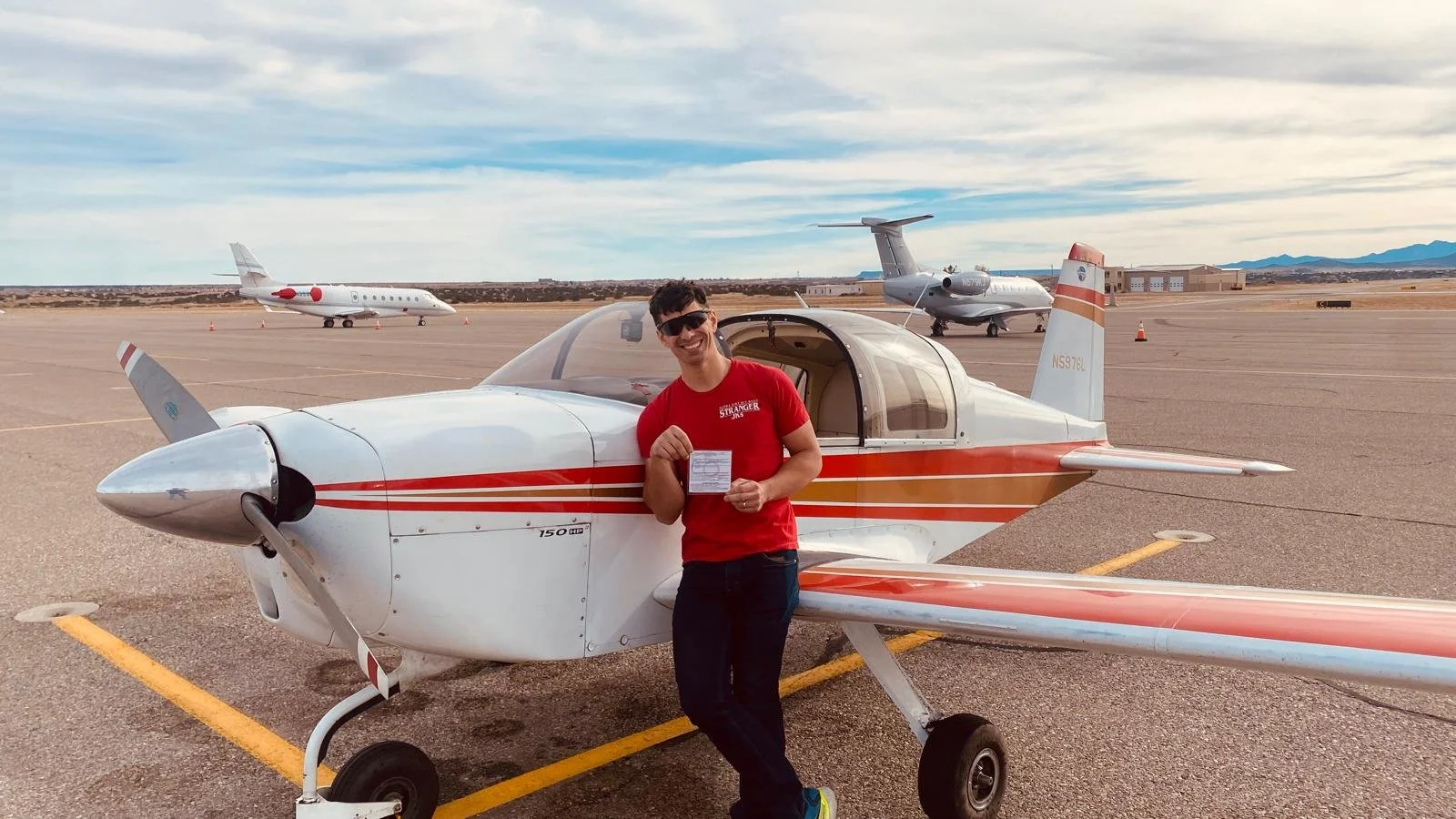 A man in sunglasses and a red shirt standing in front of a small white and red airplane on an airport tarmac, holding a boarding pass or ticket, with two larger aircraft and a hangar in the background under a partly cloudy sky.