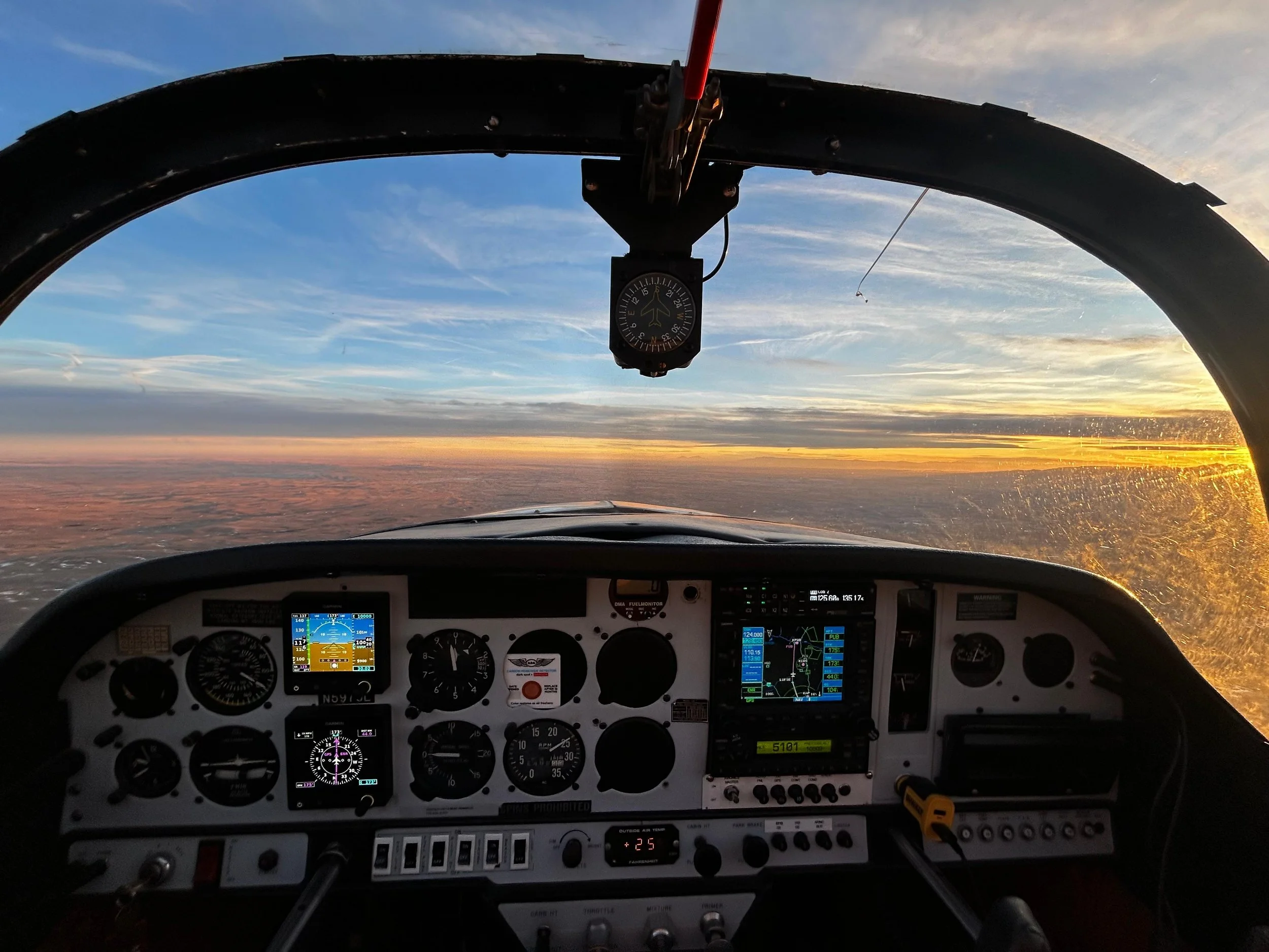 Inside view of an aircraft cockpit during sunset, showing various instruments on the dashboard and the sky with clouds and sunlight outside.