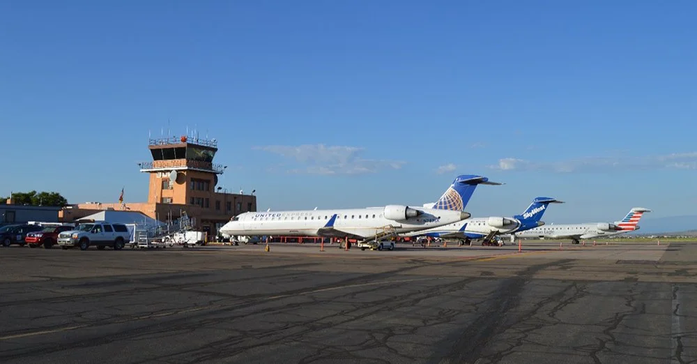 Airplanes parked at an airport gate with a control tower and vehicles nearby on tarmac under a clear blue sky.