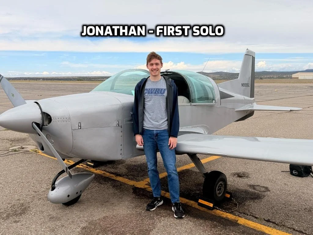 Young man standing in front of a small fighter jet aircraft on an airport tarmac, with a cloudy sky in the background. Text overlay says 'Jonathan - First Solo'.