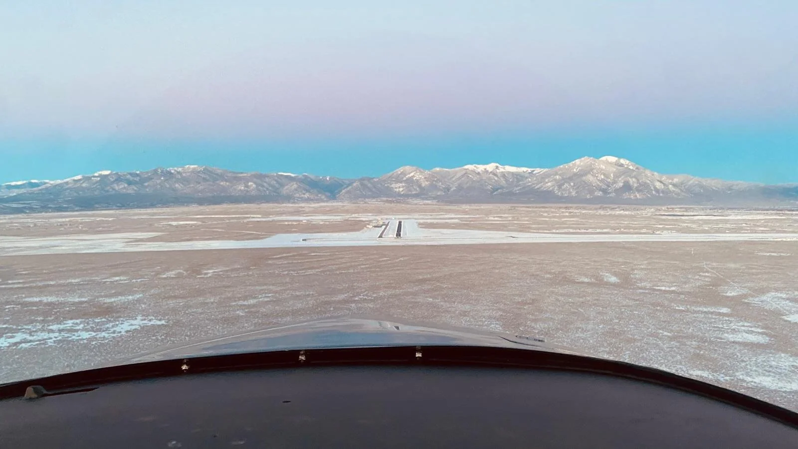 View from airplane cockpit showing runway ahead, flat snowy landscape, and snow-capped mountains under a pastel-colored sky.