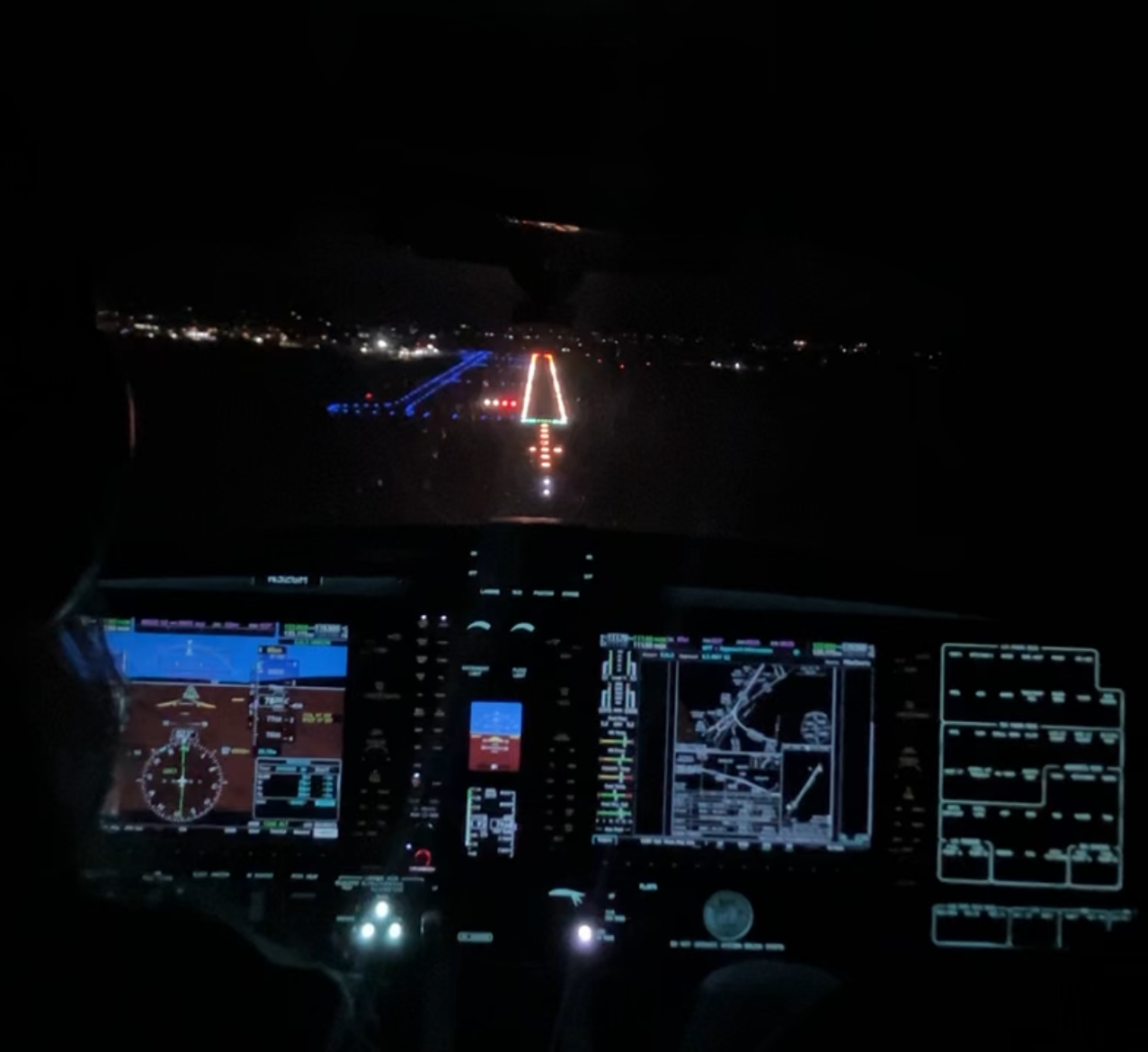 Nighttime view from the cockpit of a plane approaching a runway decorated with lights, with aircraft instruments illuminated on the dashboards.