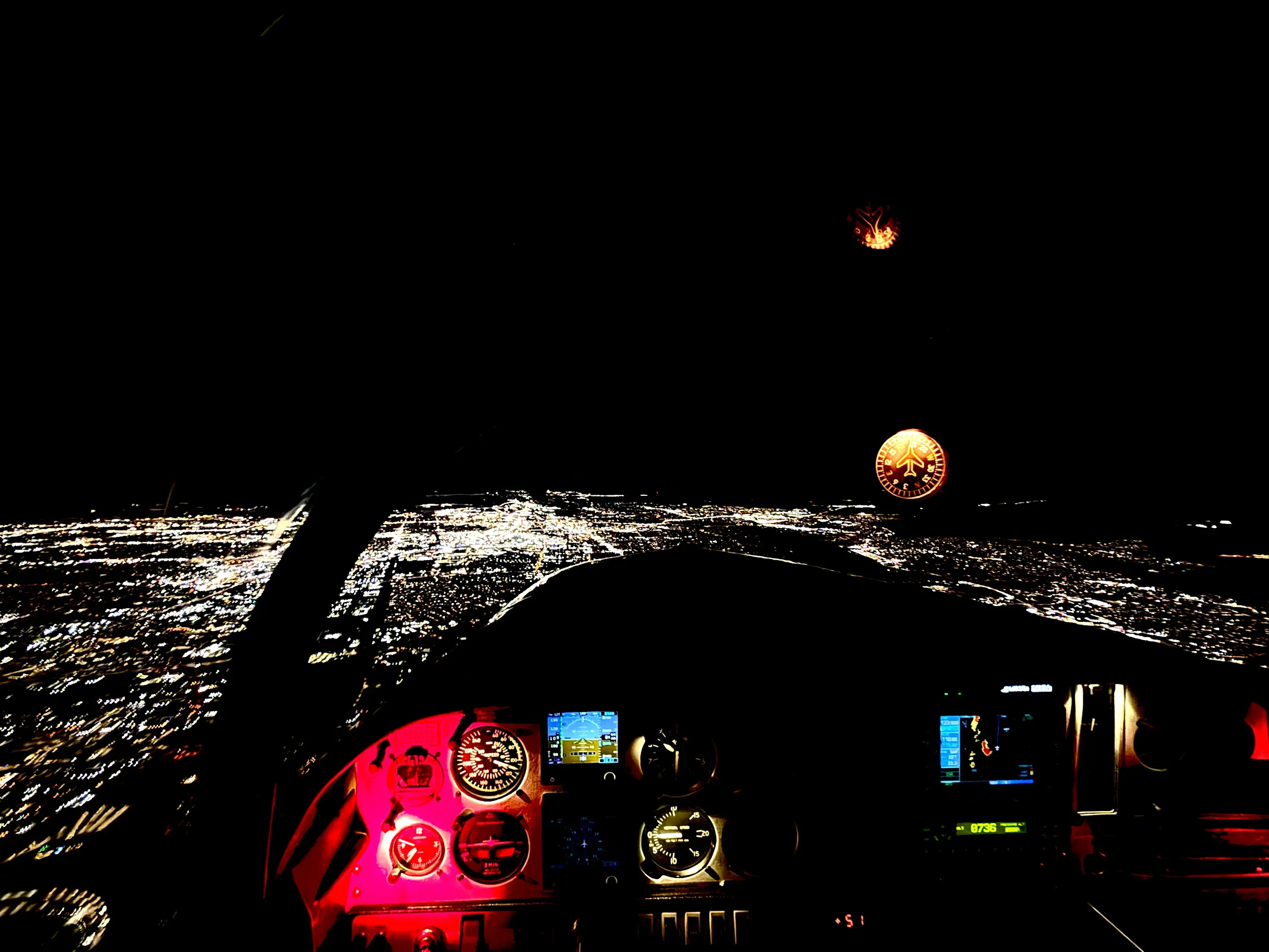 Nighttime view from inside a cockpit of an airplane flying over a city with illuminated streets, with digital flight instruments and navigation screens visible, and bright city lights below.