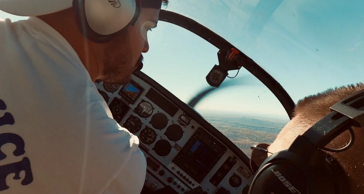 Pilot flying a airplane during daytime, view of cockpit instruments, sky, and landscape below.