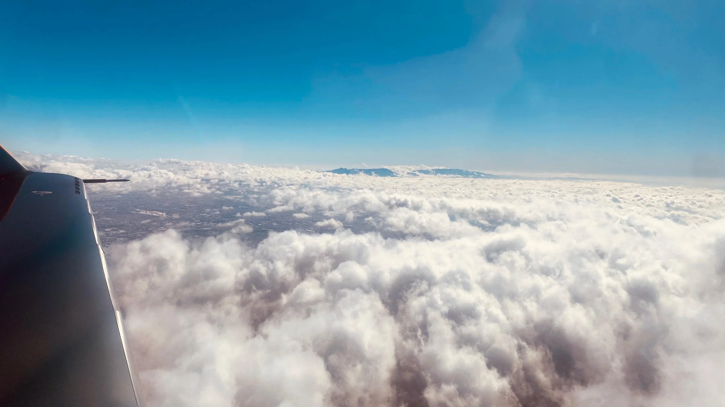View from an airplane window showing clouds below and a blue sky above with a distant landmass on the horizon.