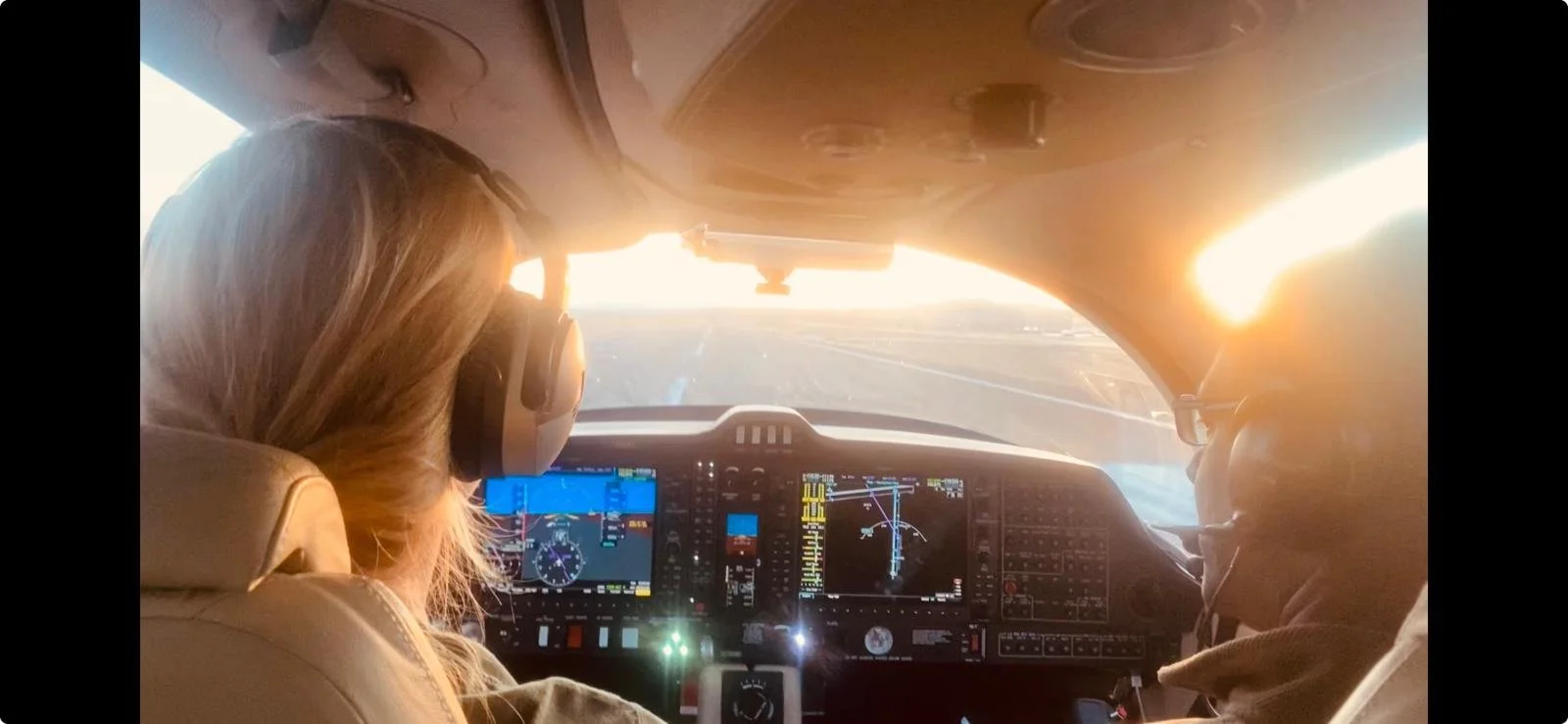 Inside an airplane cockpit with two pilots, a woman in the left seat and a man on the right, flying at sunset with the sky brightening.