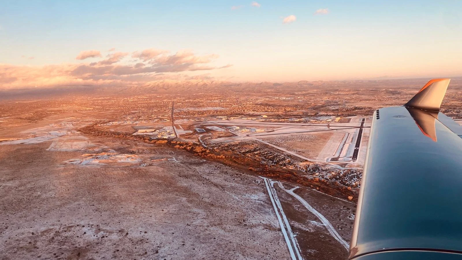 View from an airplane showing a landscape with snow patches, roads, and buildings at sunset with mountains in the background.