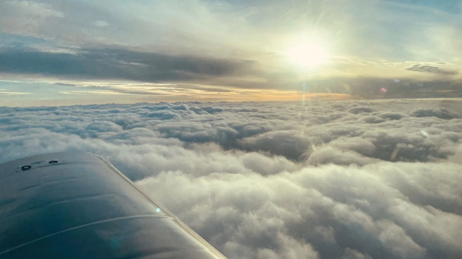 View from an airplane window showing a bright sun above clouds with a portion of the airplane wing in the bottom left corner.