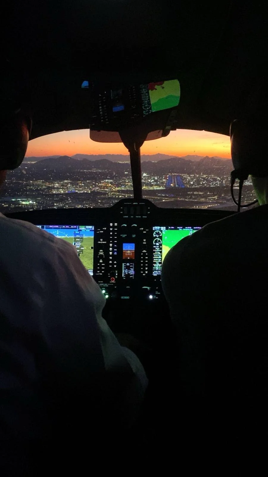 Inside the cockpit of an aircraft at dusk, showing two pilots, with illuminated control panels and a view of the city and sunset through the windshield.