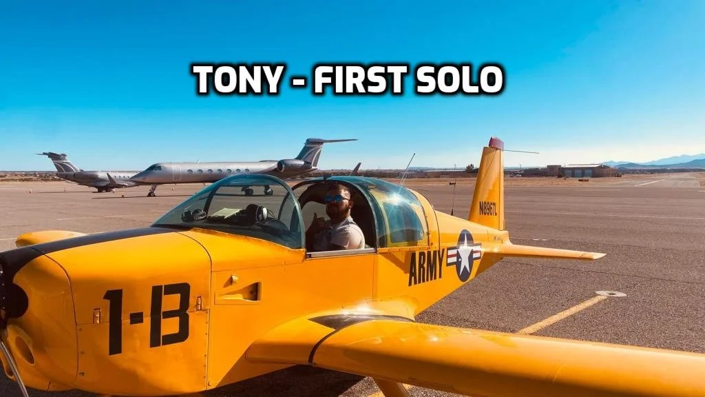 A yellow military trainer aircraft with a person sitting inside, parked on an airstrip with larger jets in the background, blue sky, and mountainous terrain in the distance. Text overlays say "TONY - FIRST SOLO."