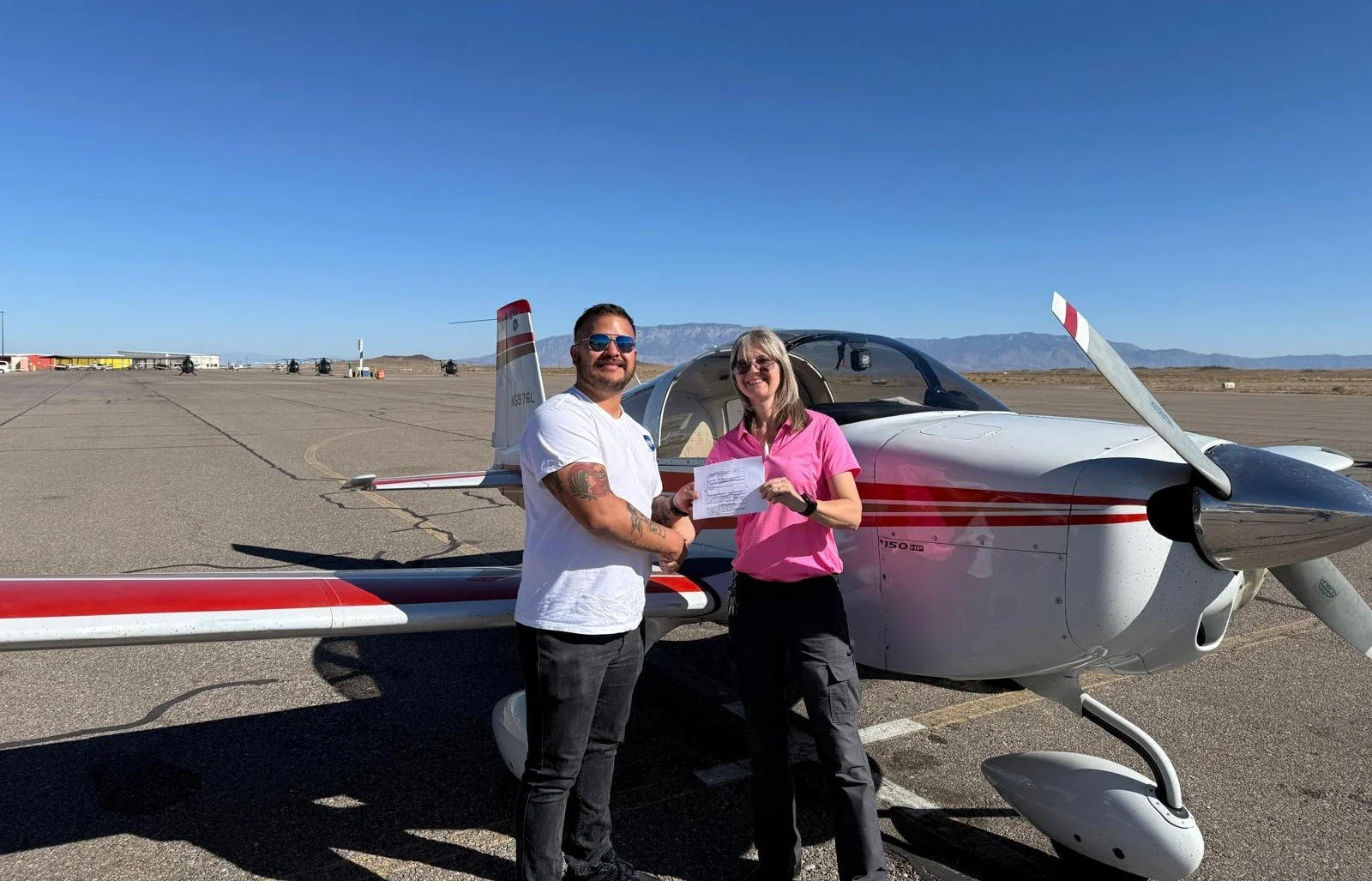 A man and a woman stand beside a small airplane on a tarmac, smiling and shaking hands, with the man holding a piece of paper. The background features a clear blue sky and distant mountains.