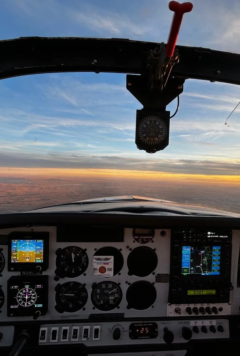 Inside the cockpit of a small aircraft flying at sunset, showing flight instruments with a view of the sky and clouds below.