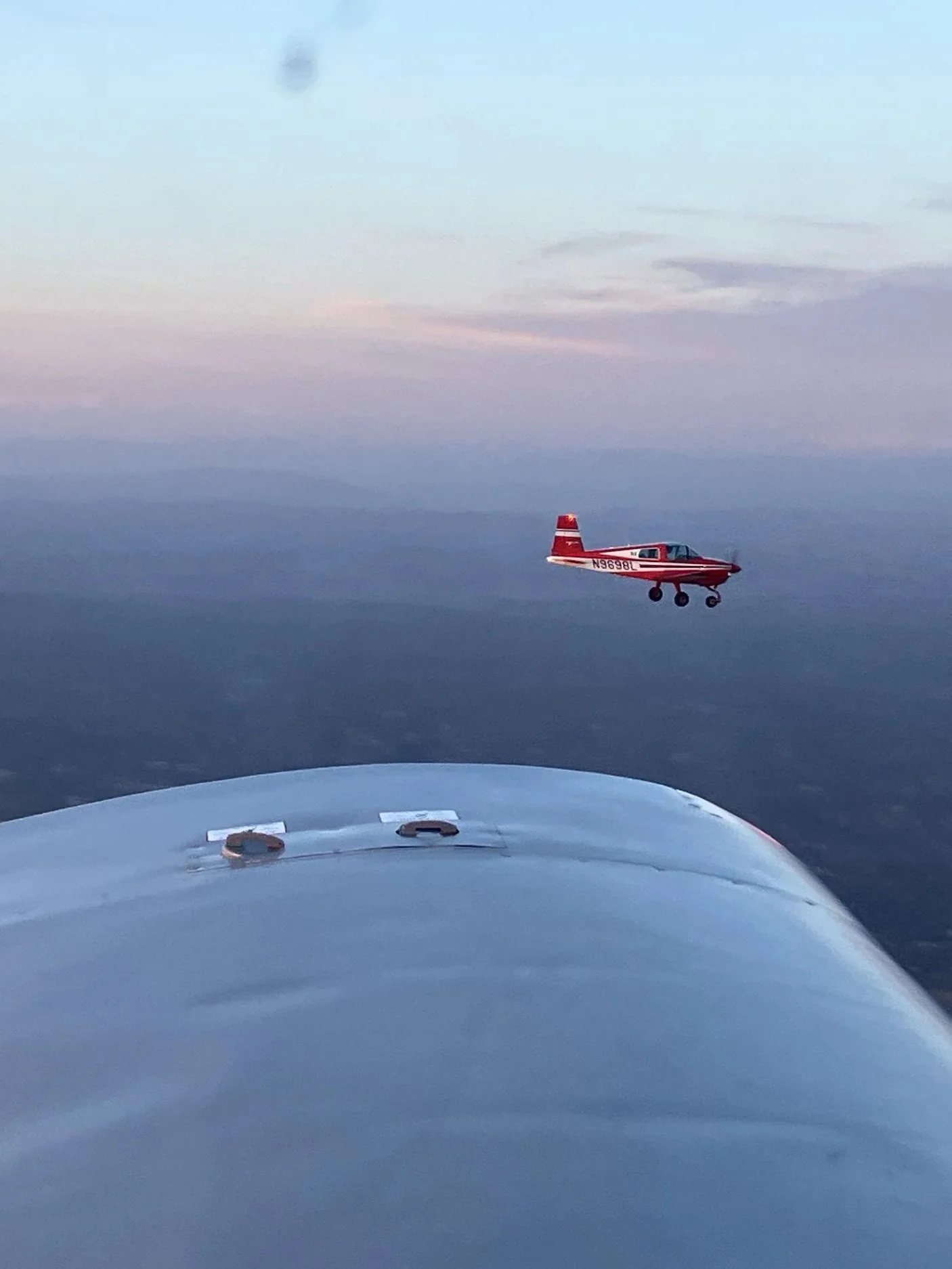 A small red and white aircraft flying above a mountain range during dusk or dawn, with the aircraft's shadow and the wing of the aircraft visible in the foreground.