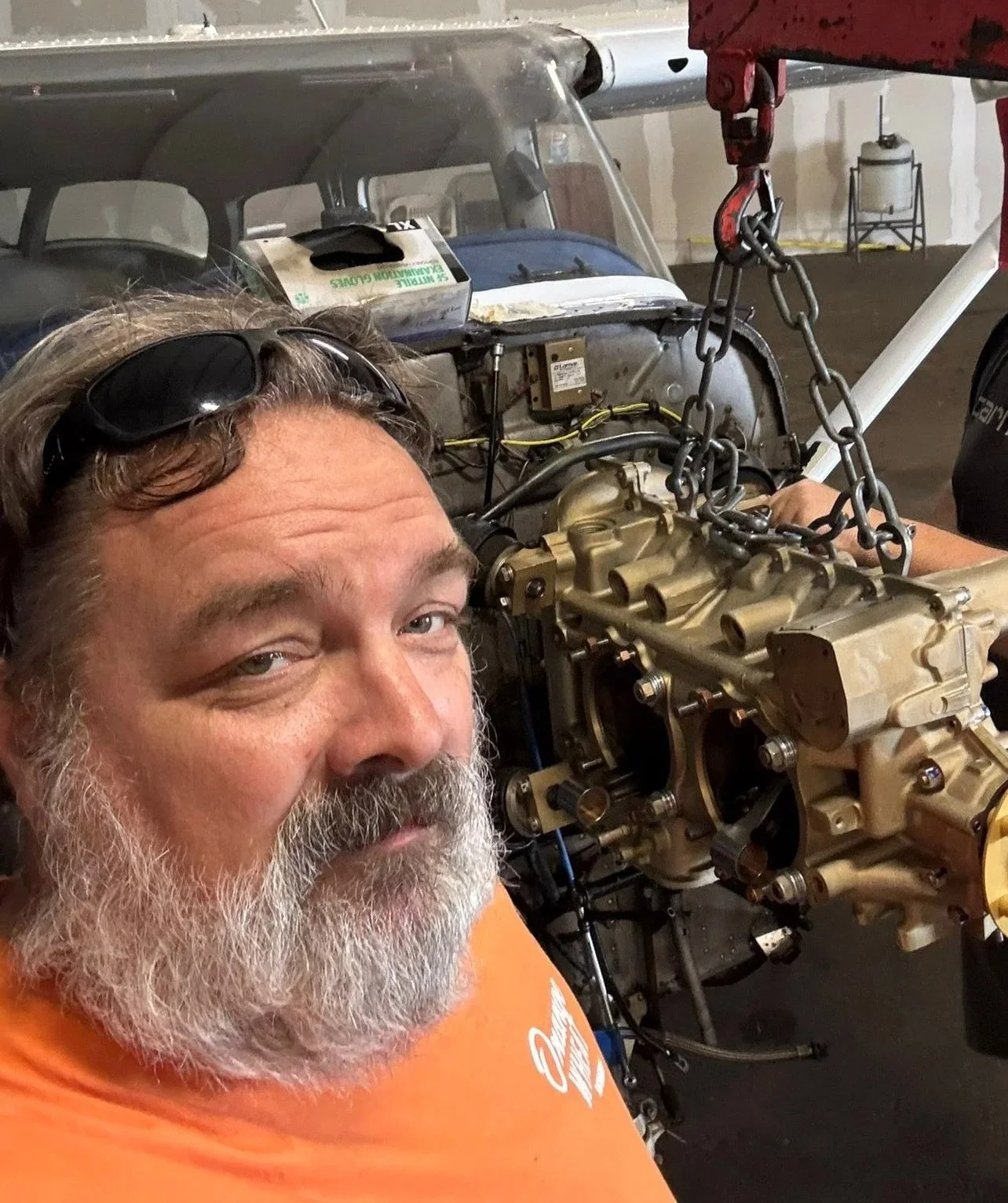 A man with sunglasses on his head and a white beard is smiling at the camera while working on a car engine in a garage.