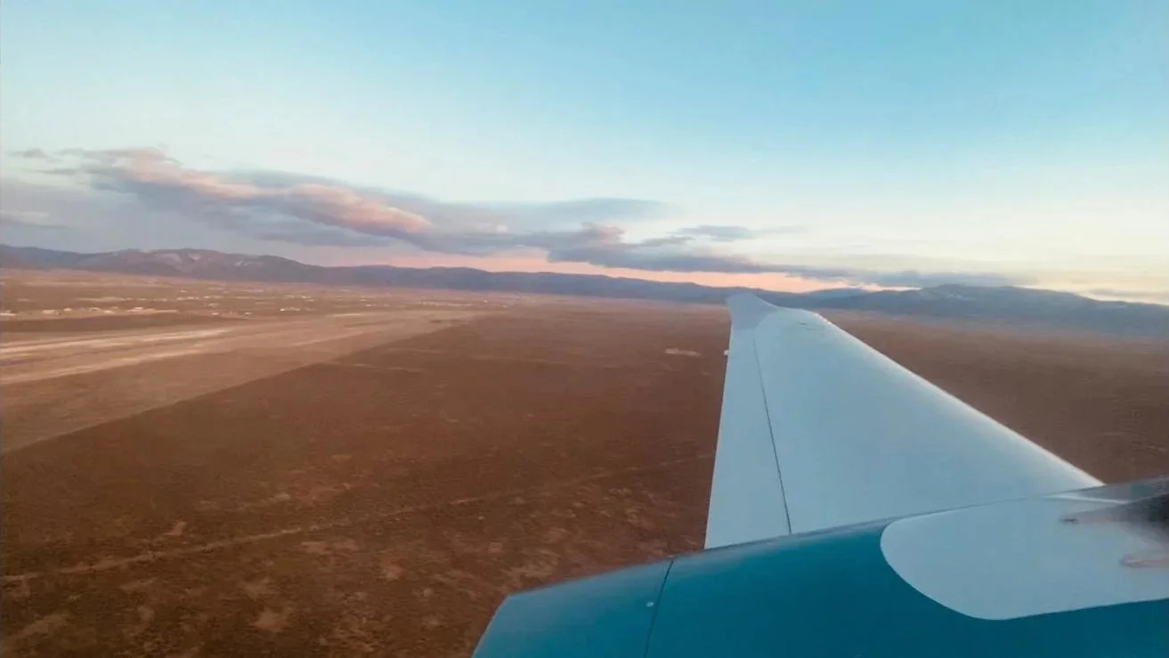 View from an airplane window showing the wing and landscape below with mountains far in the distance during sunset.