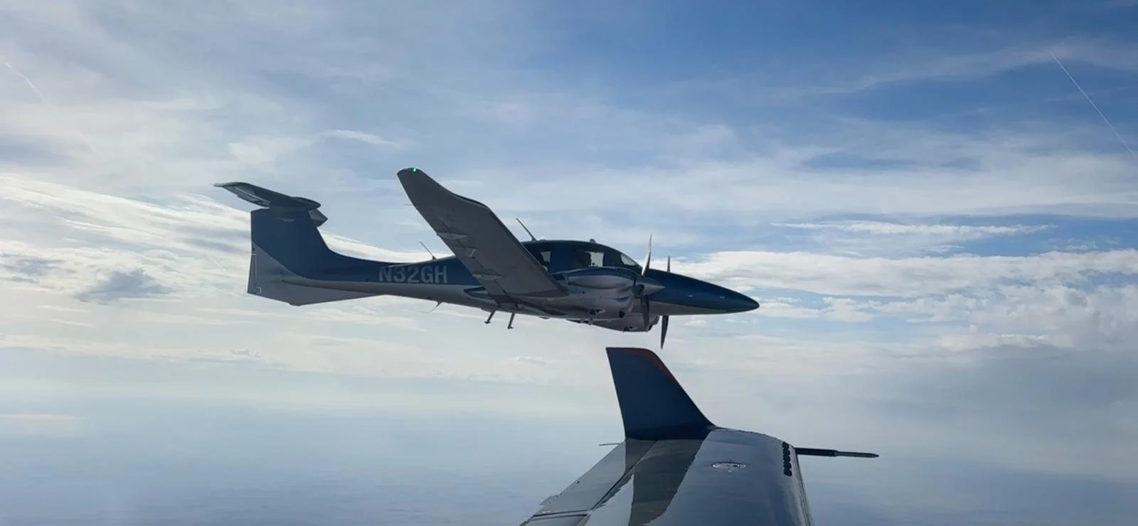 A blue and gray propeller airplane flying in the sky with clouds, seen from another aircraft.