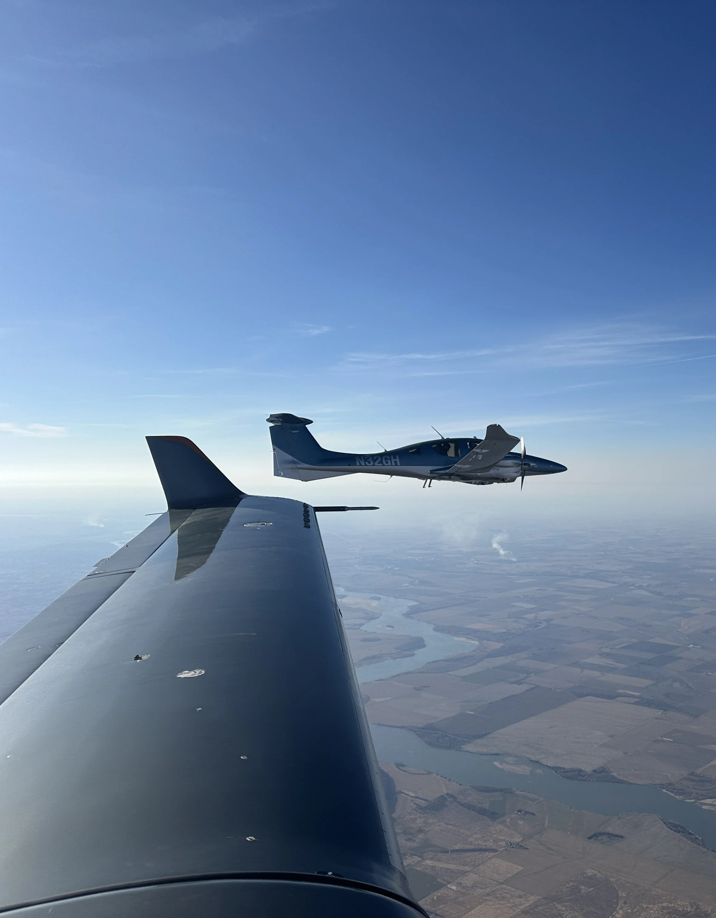 View from an airplane window showing a second airplane flying at a similar altitude, with snow-covered ground and a river below under a clear blue sky.