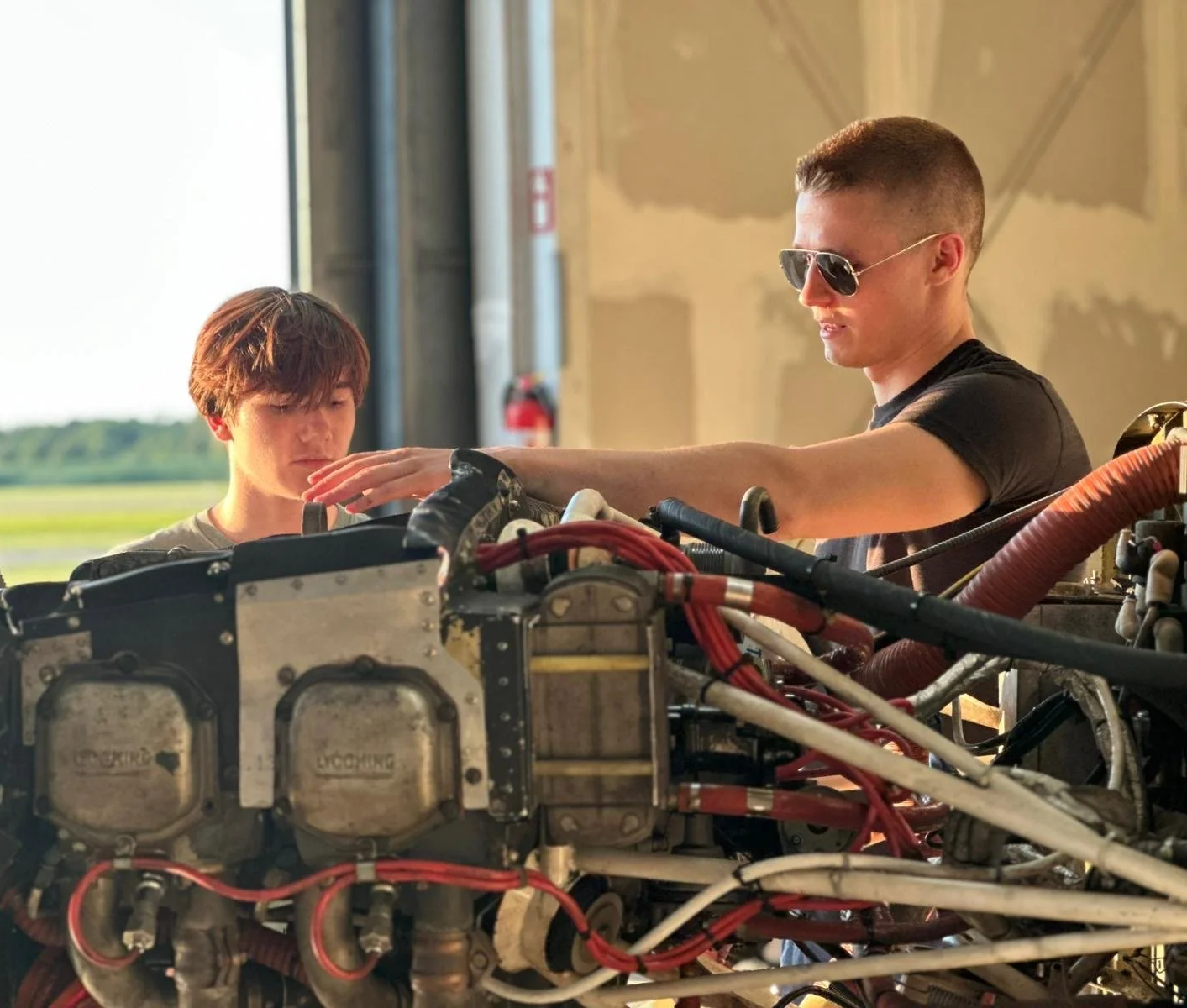 Two individuals working on a complex machine or vehicle with exposed wiring and mechanical parts inside a workshop or hangar; one person wearing sunglasses and one with brown hair.