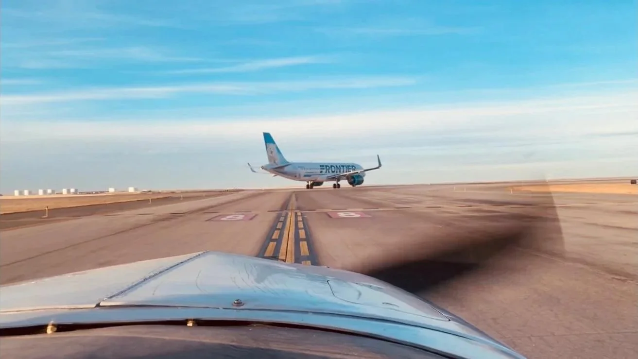 Image taken from inside a car on a runway, showing an airplane ahead with the word 'FRONTIER' on its side, preparing for takeoff or landing under a clear blue sky.
