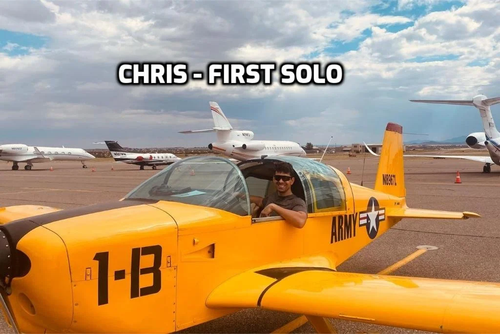 A young man sitting in a yellow military jet on an airport tarmac, with several private jets and airplanes in the background under a partly cloudy sky.