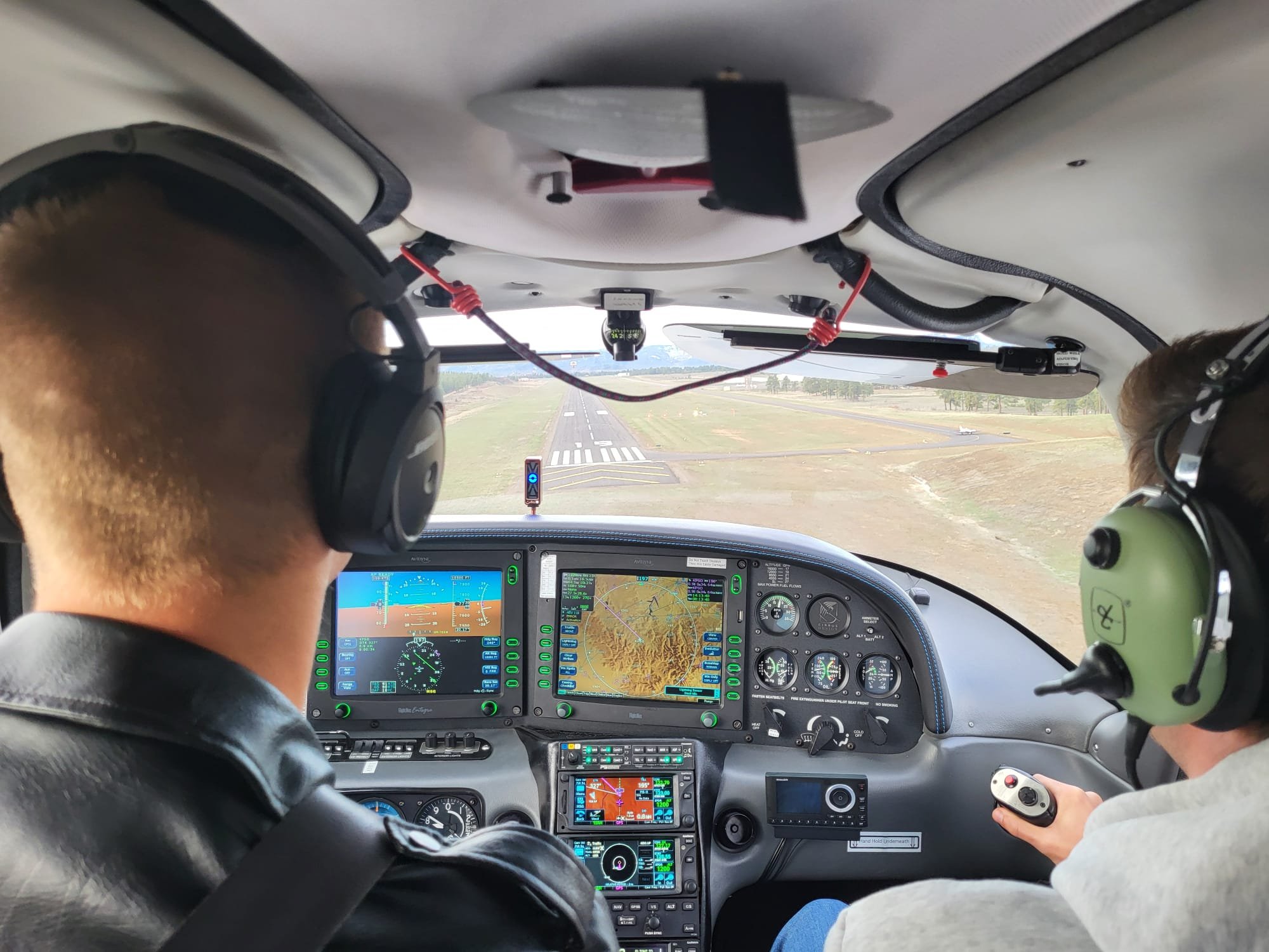 The cockpit of a small aircraft with two pilots inside. The pilot in the foreground has blond hair and wears a black jacket and headset. The co-pilot in the background wears gray clothing and holds a control device. The aircraft is flying over a runway with open fields and a plane visible in the distance.