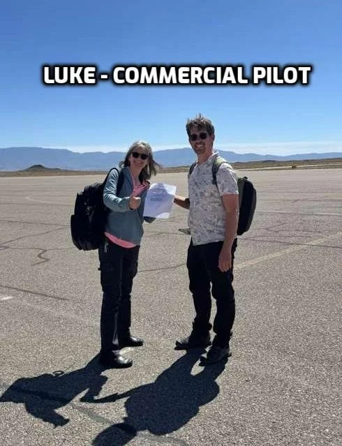 Two people, a woman and a man, standing on an airport tarmac, holding a paper, with backpacks. The woman is wearing sunglasses and a jacket, and the man is wearing sunglasses and a short-sleeve shirt. The background shows mountains and a clear blue s