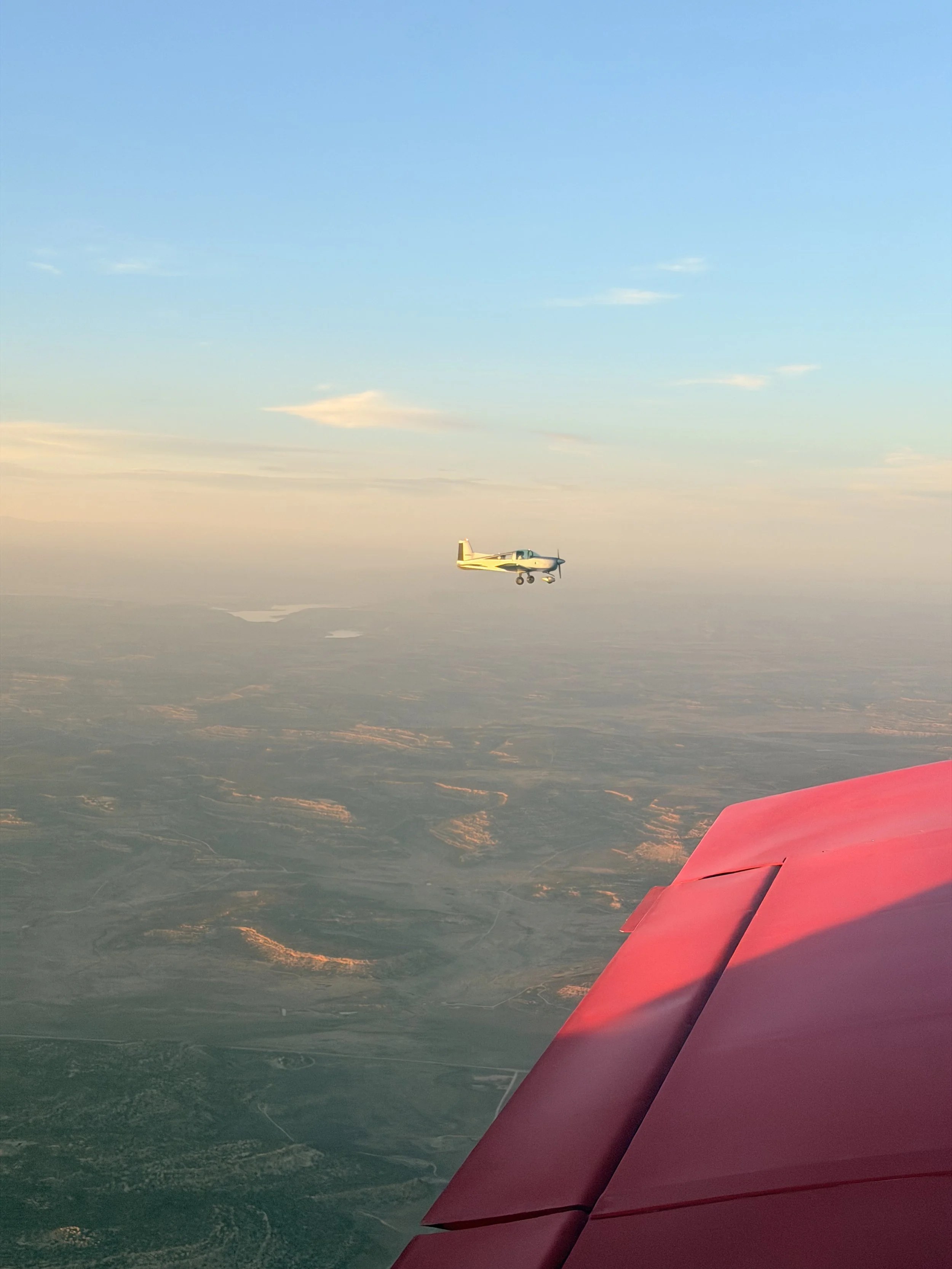 A small plane flying above a landscape during sunset with a pink aircraft wing visible in the foreground.