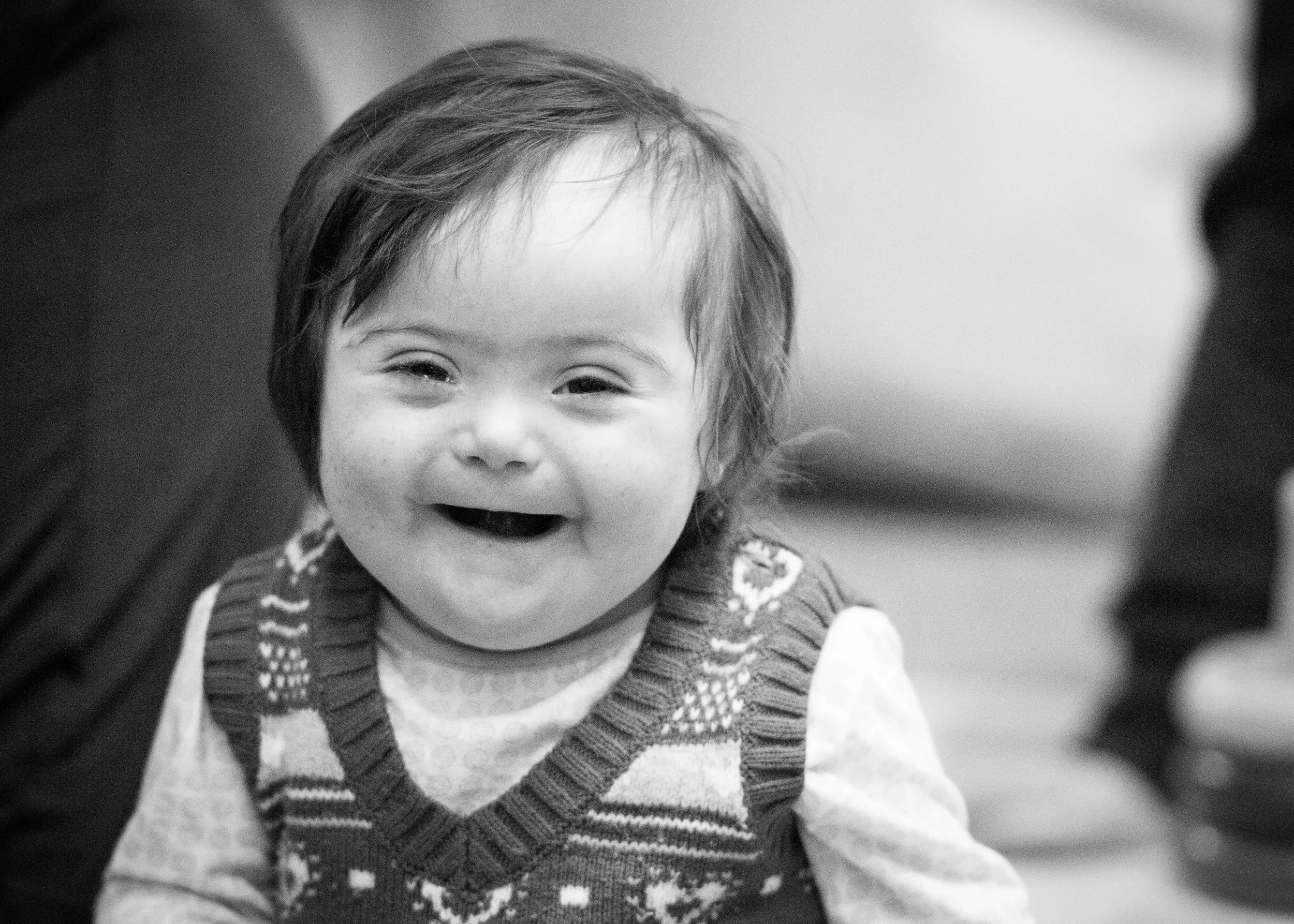 Black and white photo of a smiling young child with short hair, wearing a patterned sweater vest.