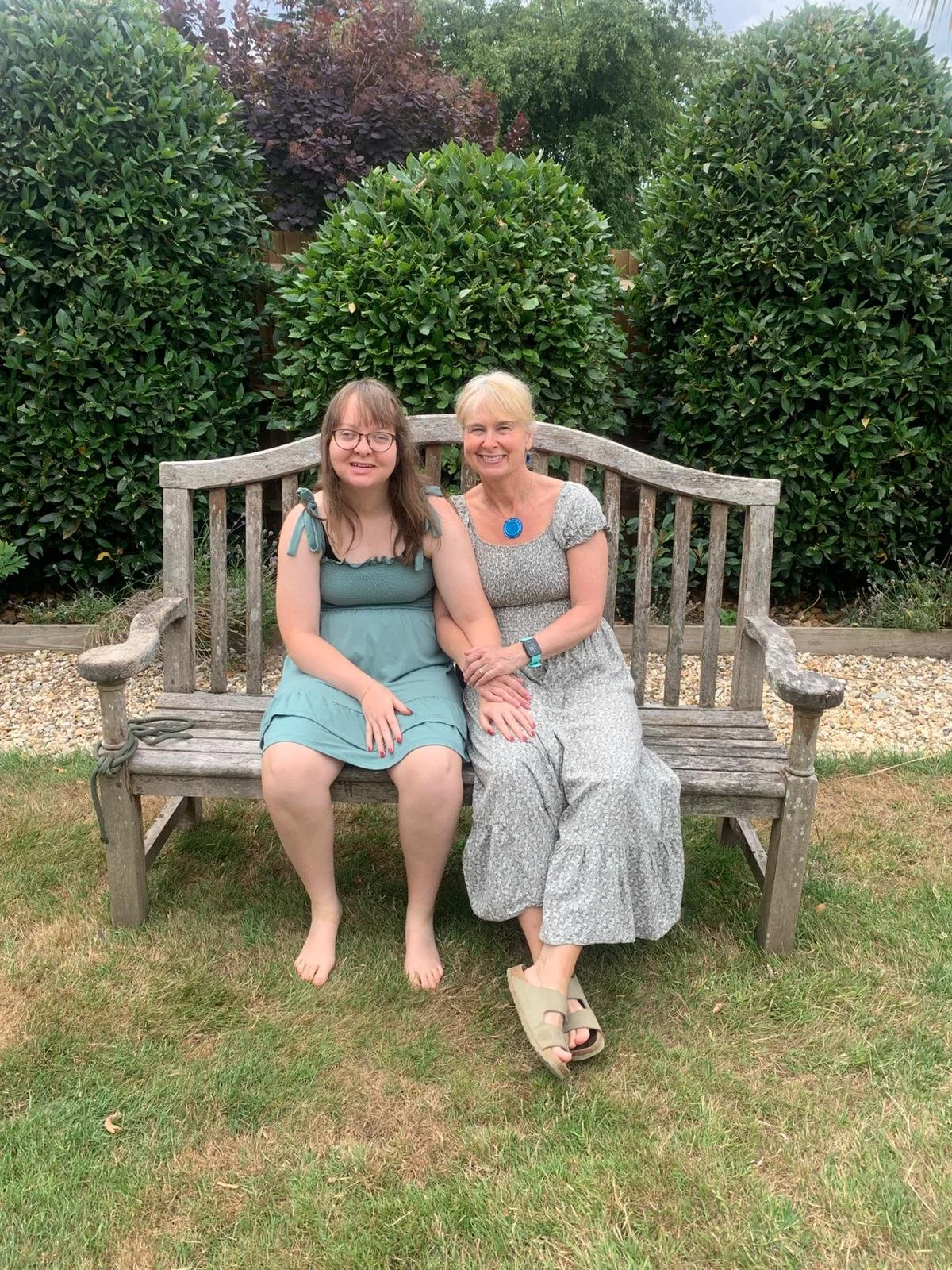 Celia and Maia founders of Challenge Group Muswell Hill sitting on a weathered wooden garden bench, smiling, with large green bushes and trees behind them.